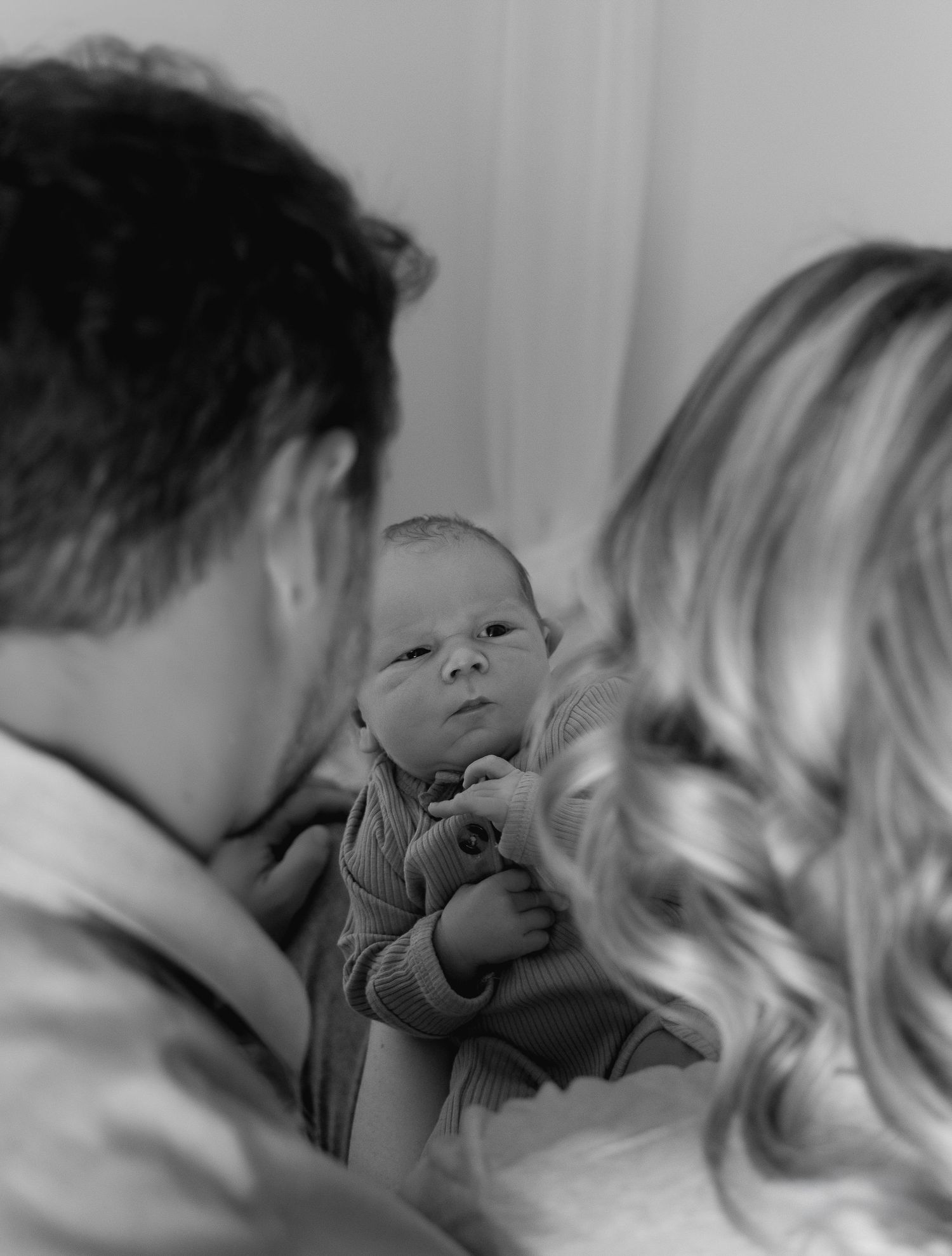 Black and white photo of two parents lovingly gazing at their newborn baby held between them.