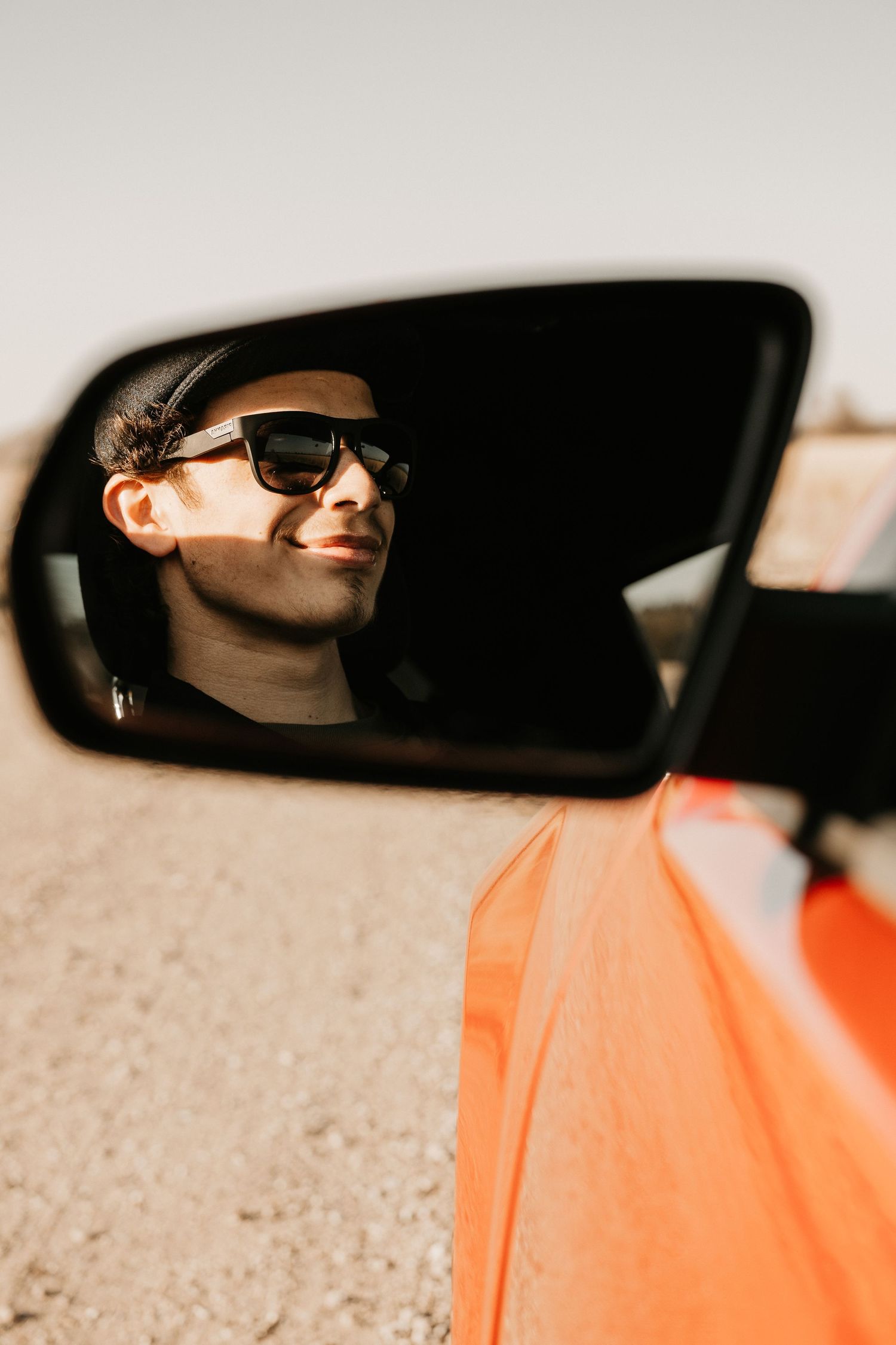 Man in sunglasses and cap reflected in orange car side mirror on a sunny desert road.