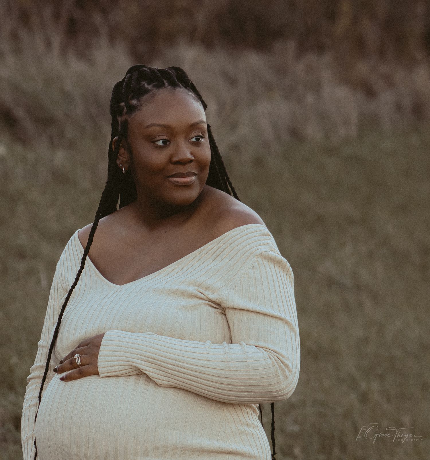 Pregnant woman in cream off-shoulder ribbed sweater poses outdoors in a field, cradling her baby bump.