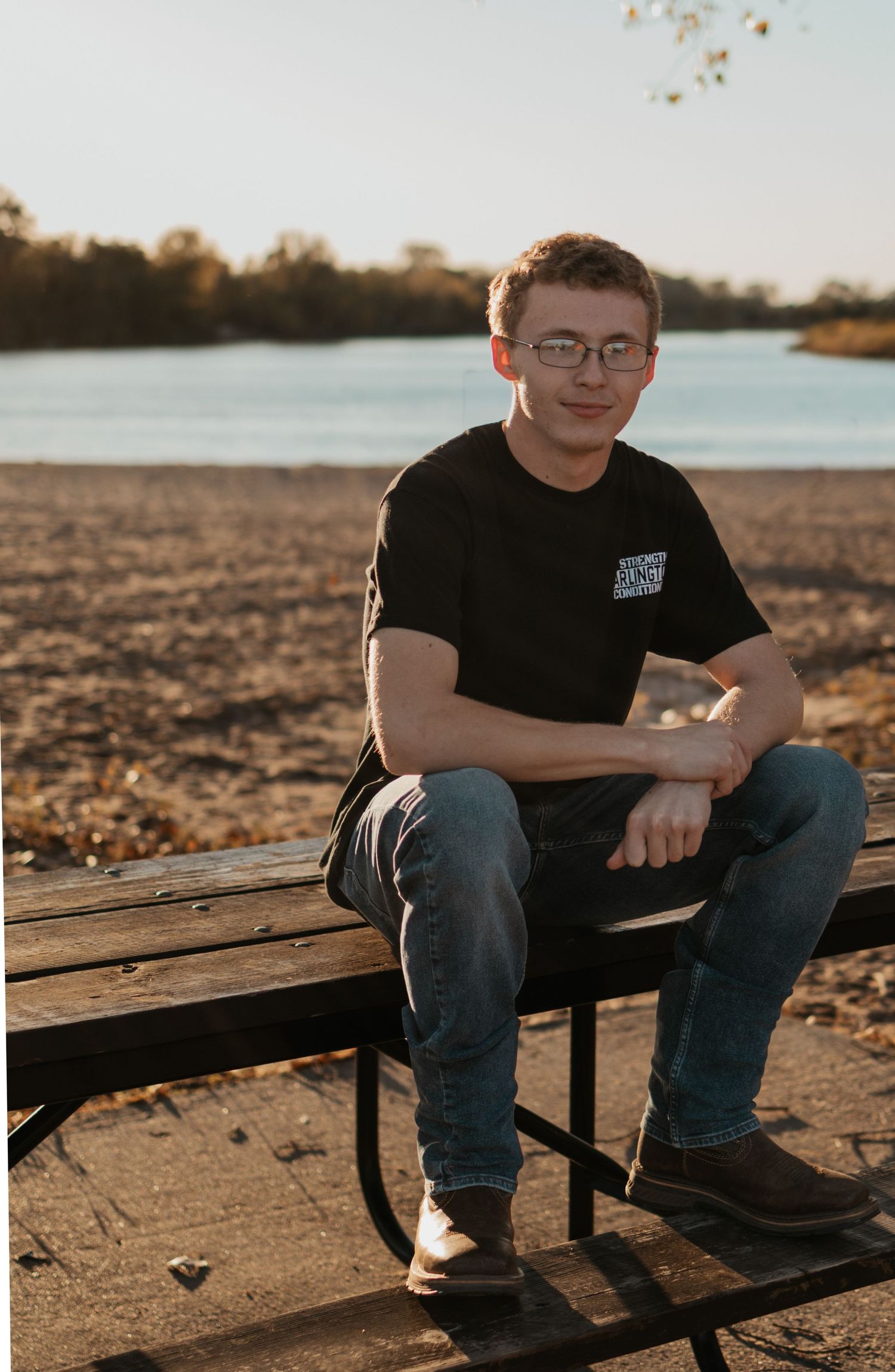 Young man in black t-shirt and jeans sitting on a picnic bench by a serene lake during golden hour.