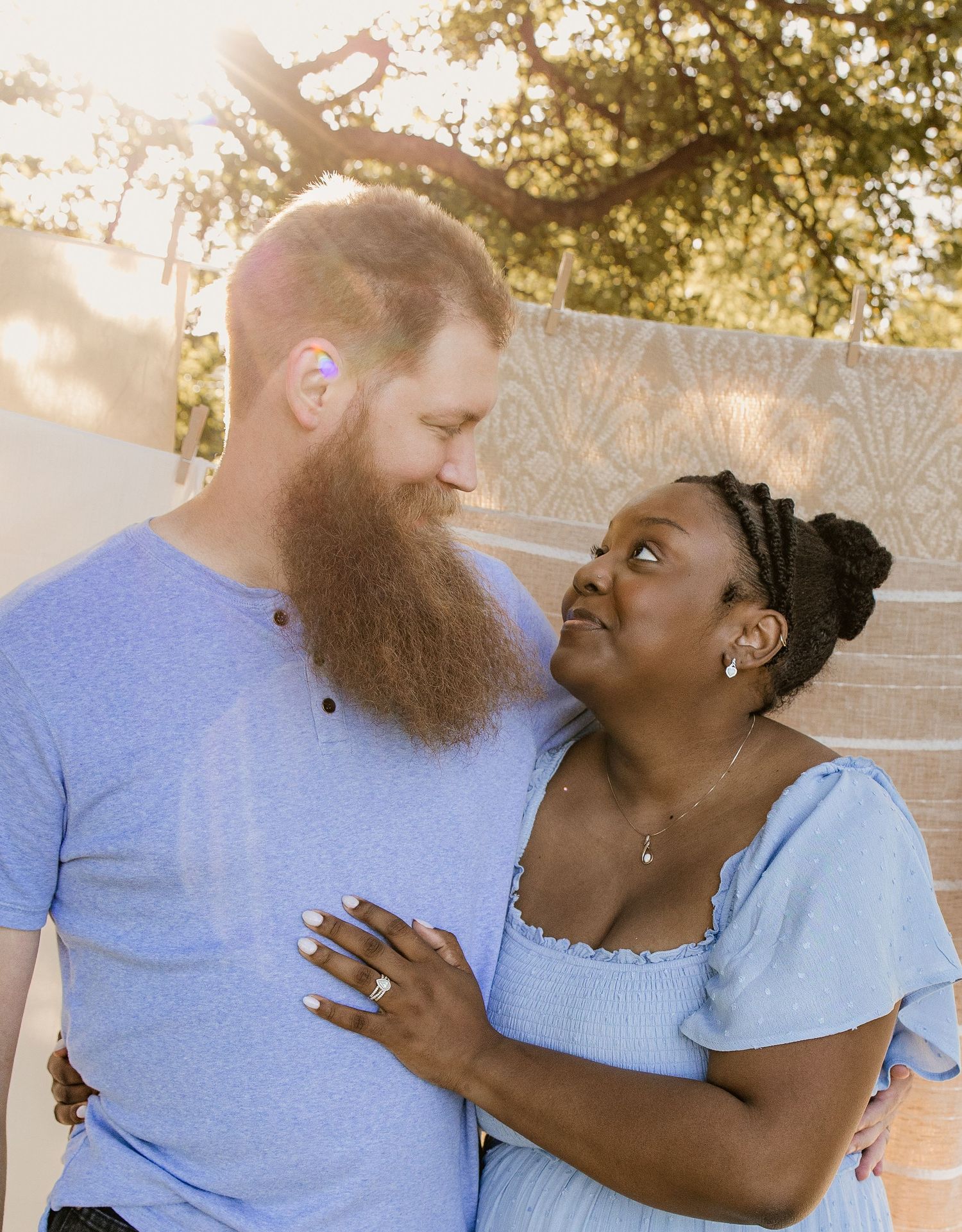Interracial couple sharing a loving gaze outdoors in golden sunlight with macrame backdrop, woman in blue dress.