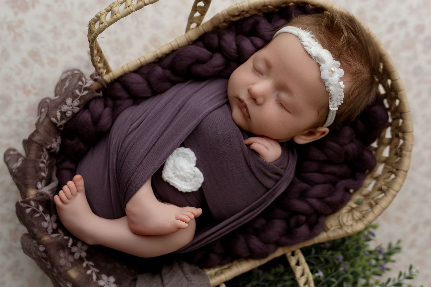 Peaceful newborn baby wrapped in purple swaddle with white floral headband resting in wicker basket.