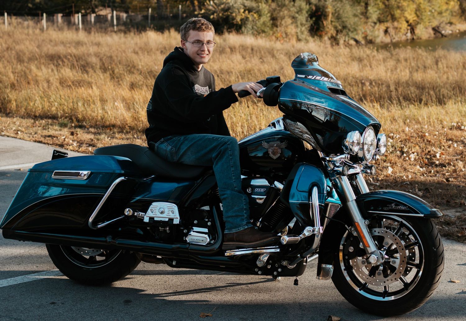 Man riding a teal Harley-Davidson motorcycle on a road with autumn trees in the background.