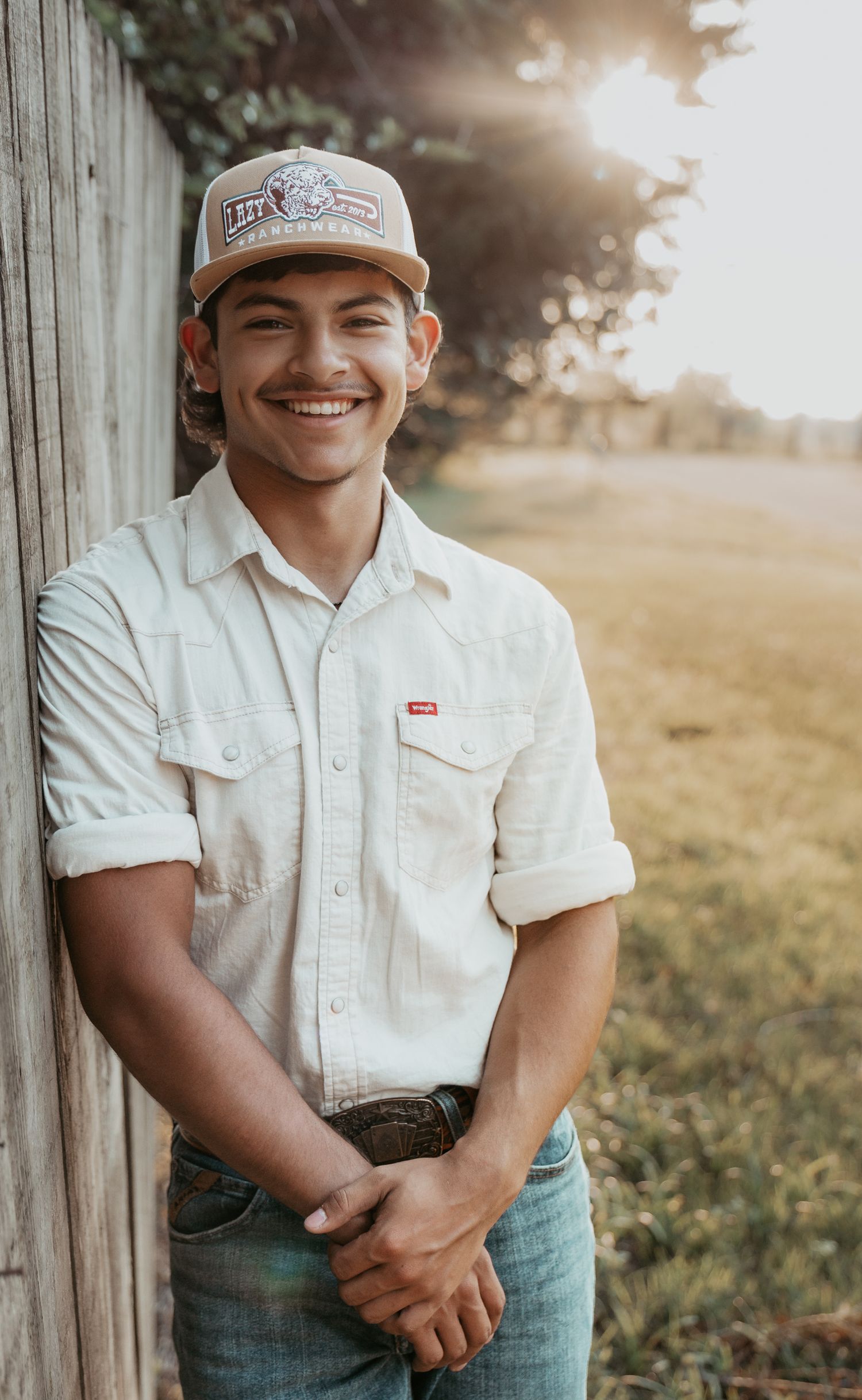 Young man in Levi's shirt and trucker hat smiling outdoors by wooden fence in golden sunlight.
