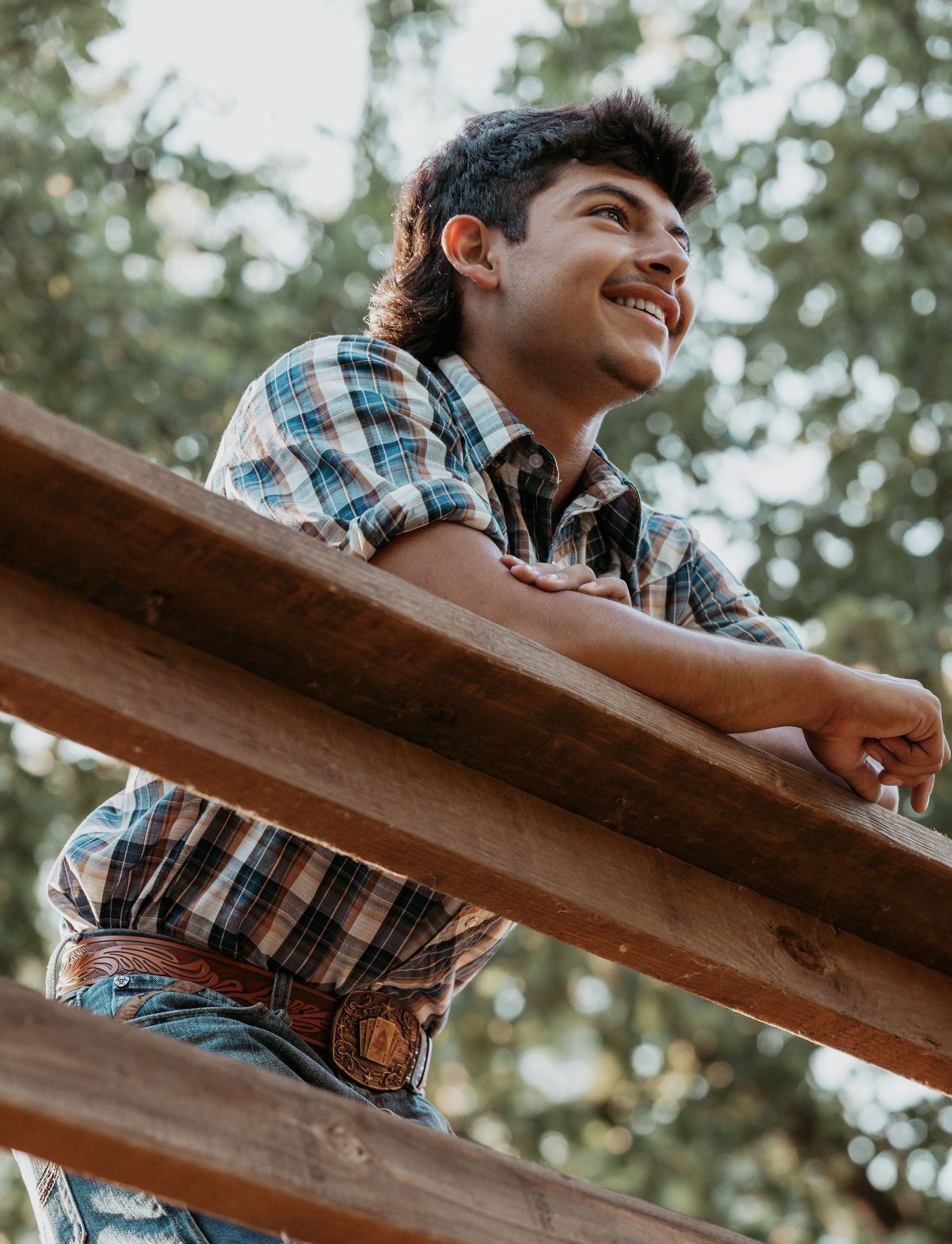 Smiling young man in plaid shirt and belt buckle leaning on wooden fence rail outdoors.