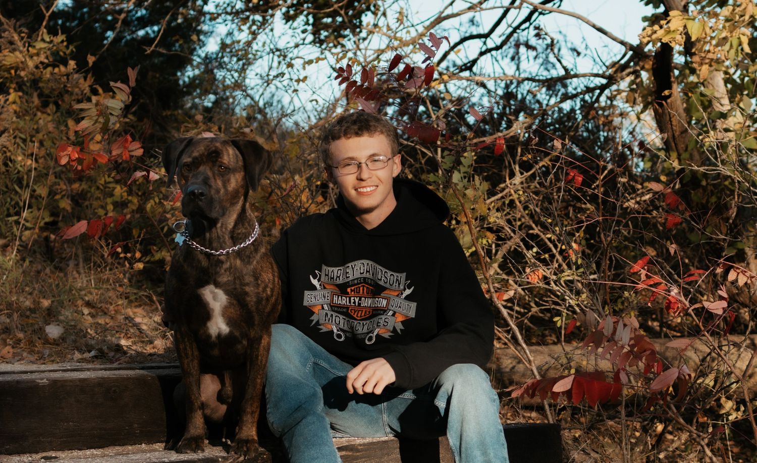 Teen boy wearing Harley-Davidson hoodie sitting outdoors with brown dog amid fall foliage.