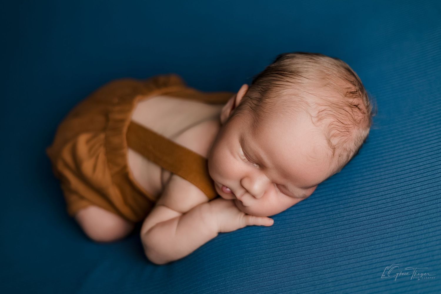 Newborn baby sleeping peacefully on blue fabric wearing brown overalls in a professional photography portrait.