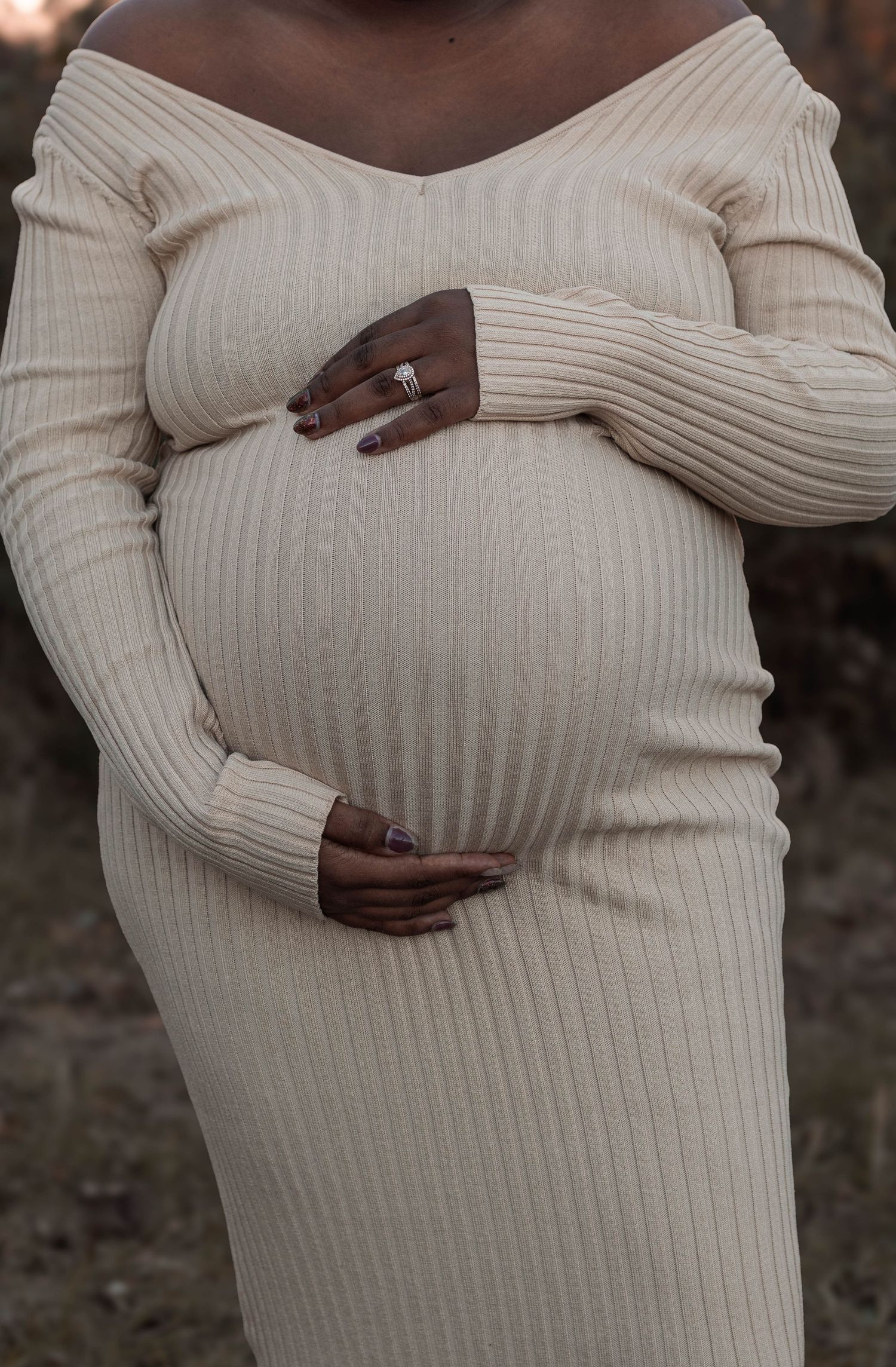 Pregnant woman in cream ribbed off-shoulder dress cradling baby bump, wearing engagement ring, maternity photo.