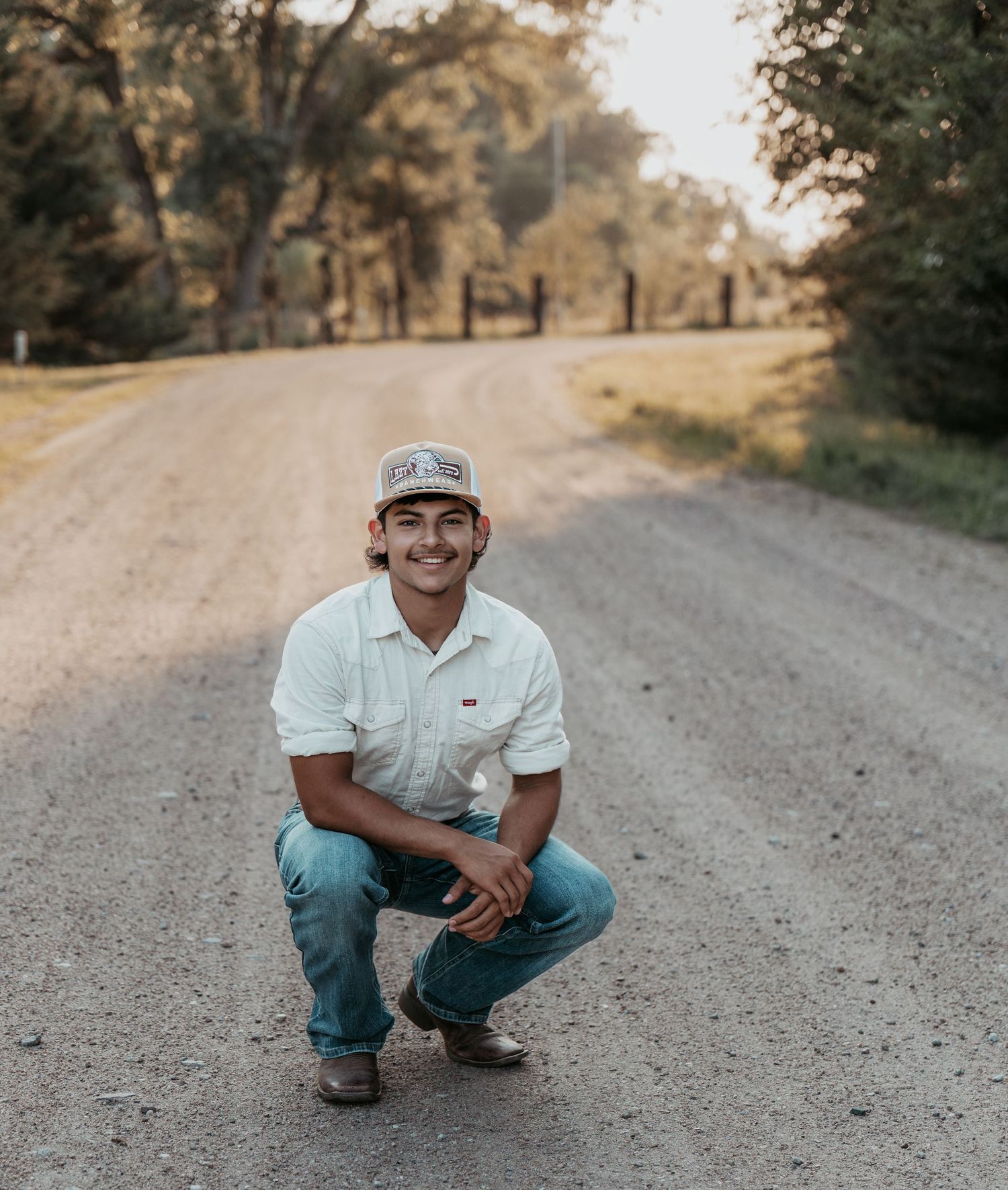 Young man in white shirt and cap crouching on a rural gravel road surrounded by trees at golden hour.