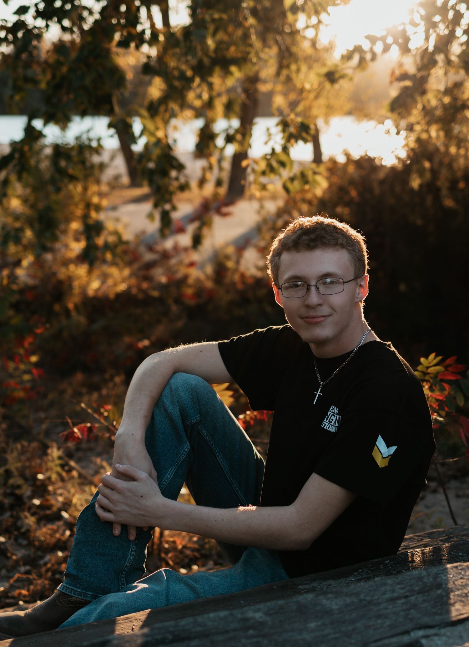 Young man in black t-shirt and jeans sitting outdoors on a log surrounded by autumn foliage near a lake.