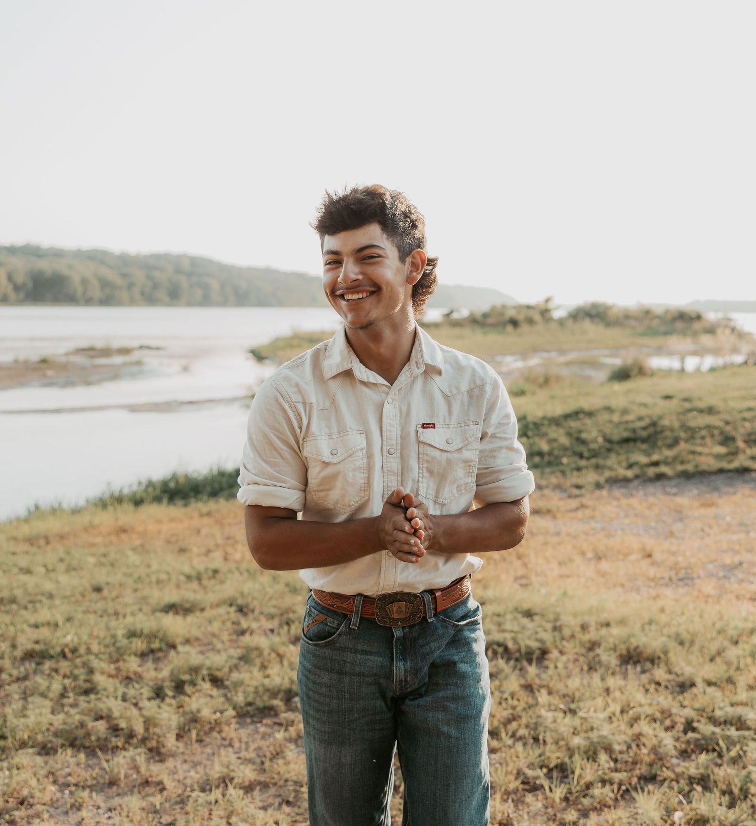 Smiling young man in white shirt and jeans standing outdoors near a river with green hills in background.