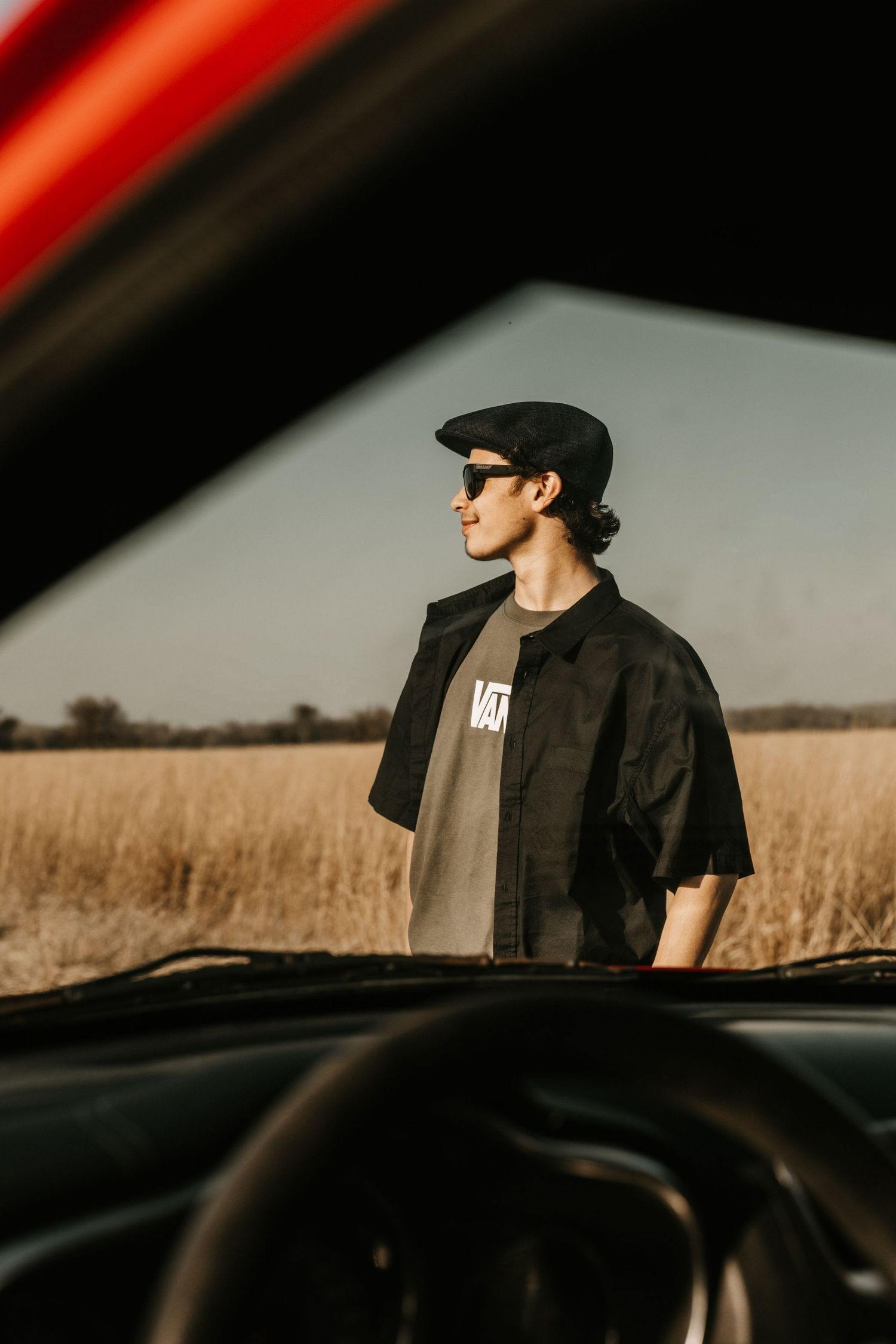 Young man in Vans shirt and flat cap standing by car in golden field.