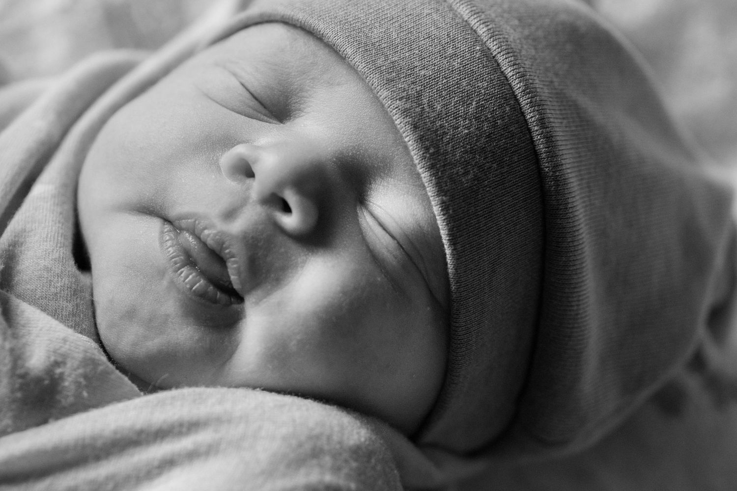Black and white close-up photo of a sleeping newborn baby wrapped in a soft blanket wearing a knit hat.