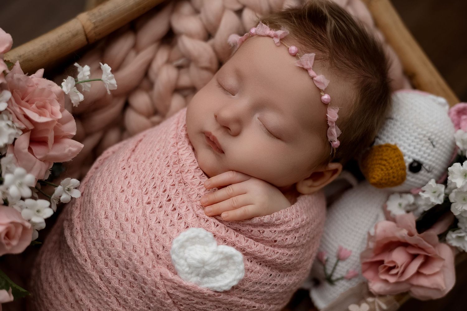 Sleeping newborn baby girl swaddled in pink knit wrap with floral headband in a basket with roses.