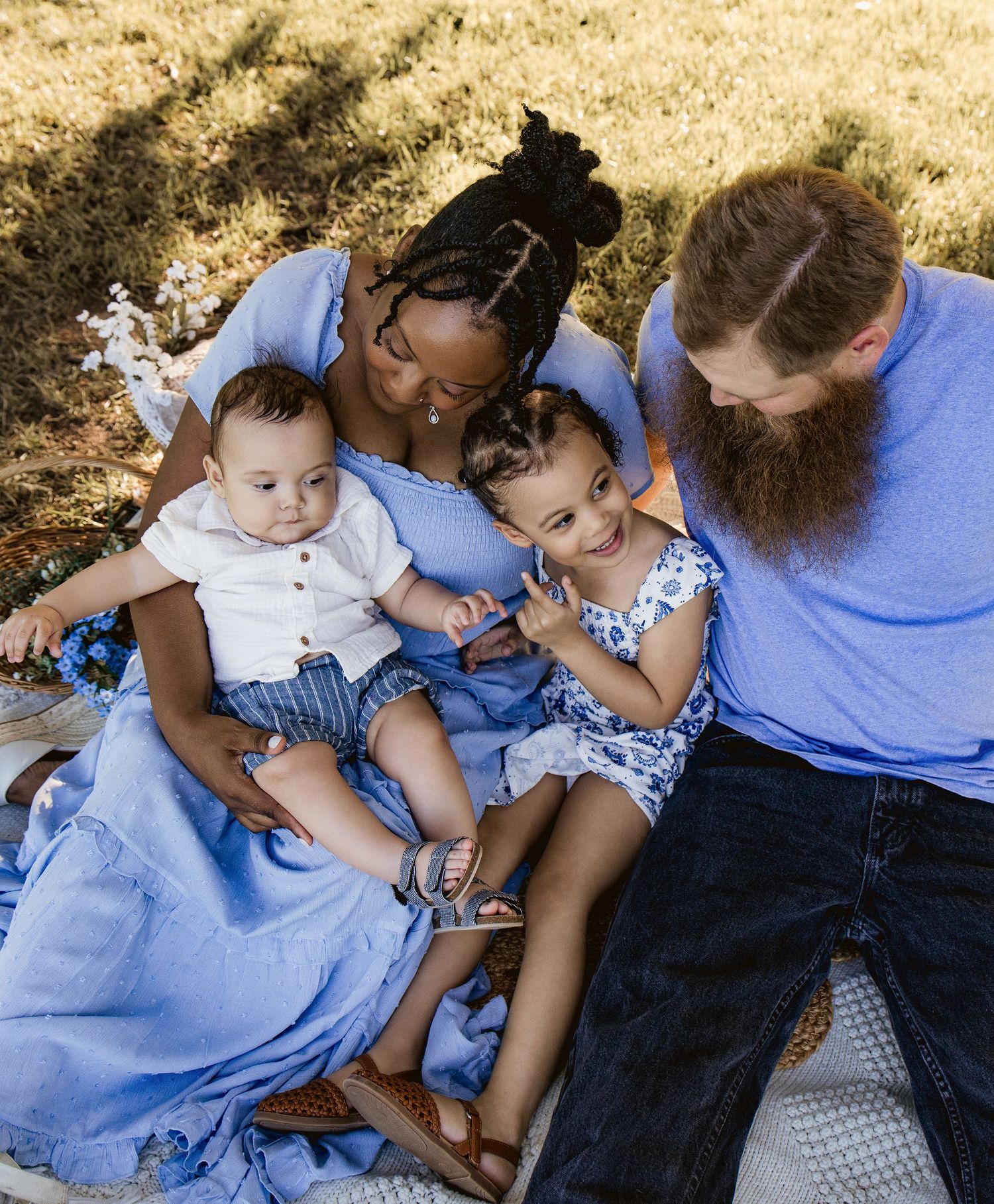 A happy interracial family of four sits on a blanket outdoors, dressed in coordinating blue outfits.