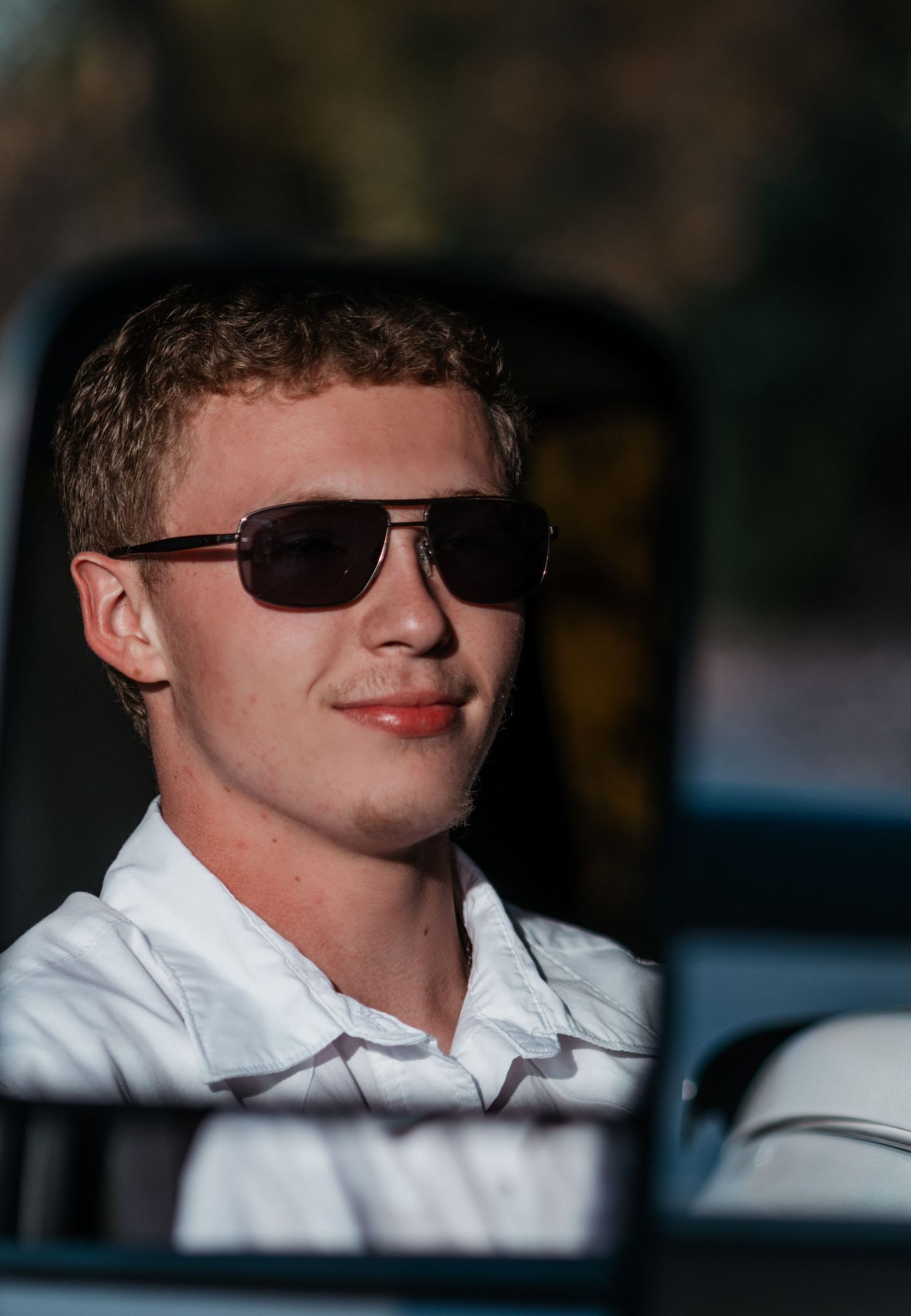 Young man wearing aviator sunglasses and white shirt sitting in a vehicle, smiling in natural sunlight.