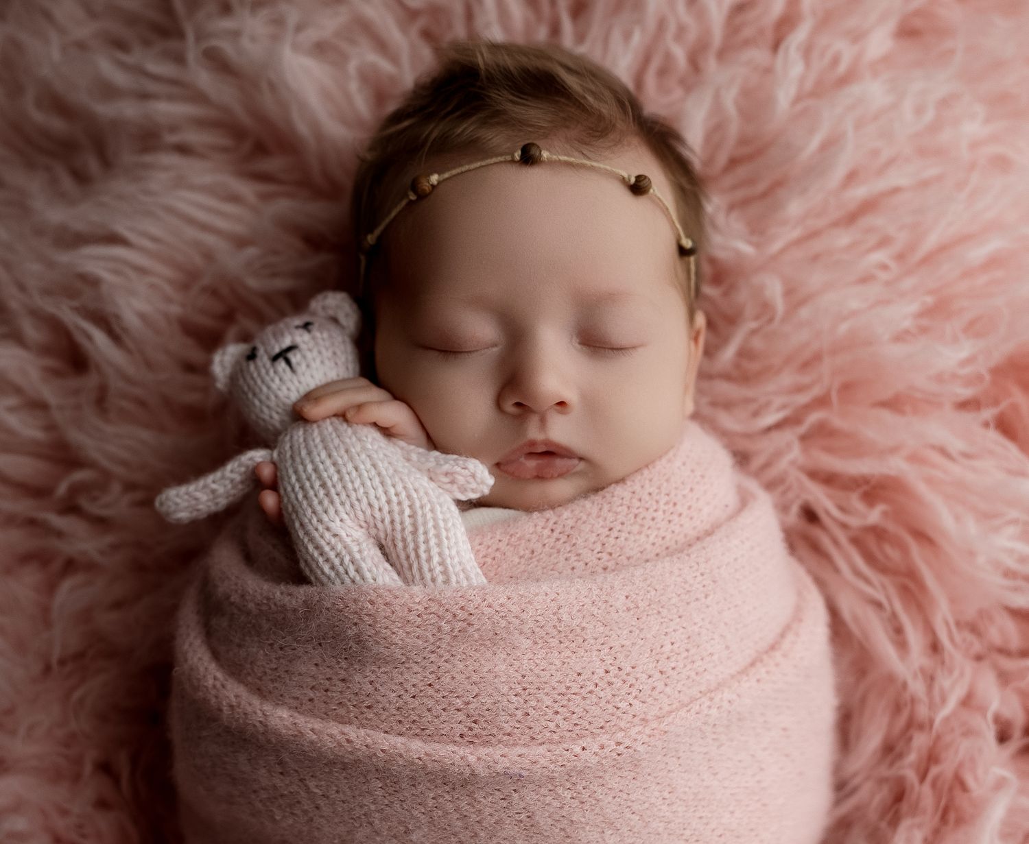Newborn baby girl swaddled in pink wrap holding knit bear toy, sleeping on fluffy pink fur backdrop.