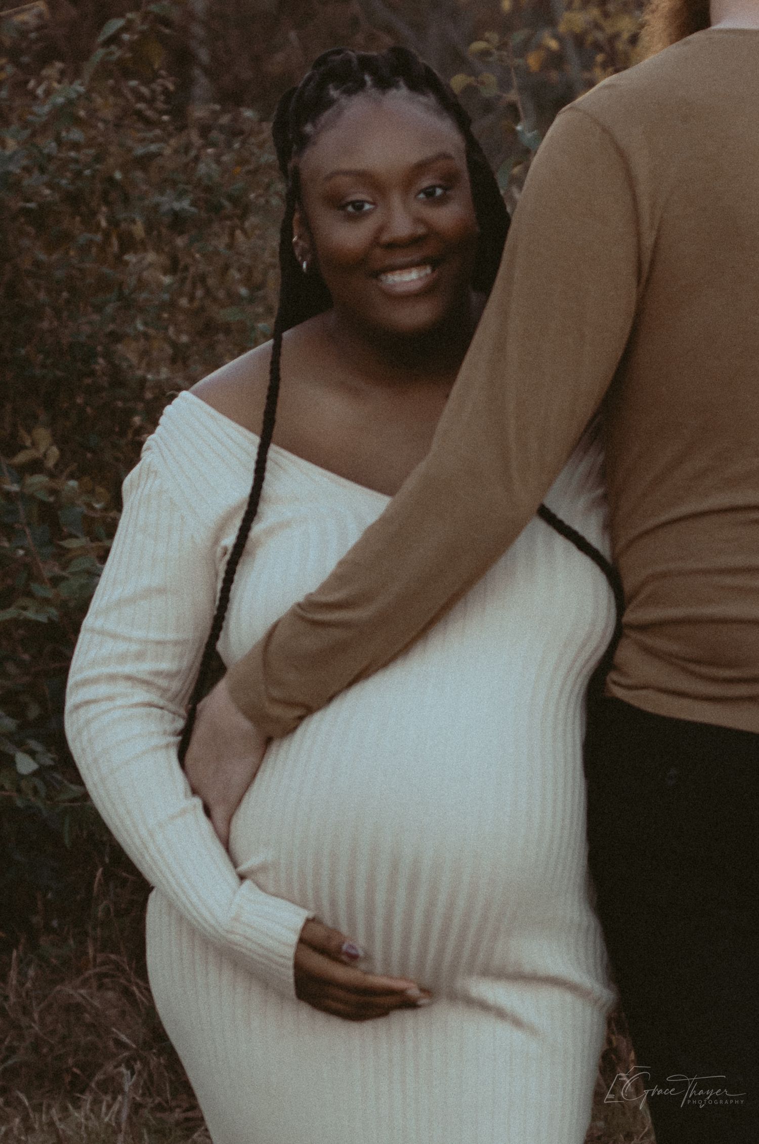 Smiling pregnant woman in white ribbed off-shoulder dress poses outdoors with partner during maternity photoshoot.