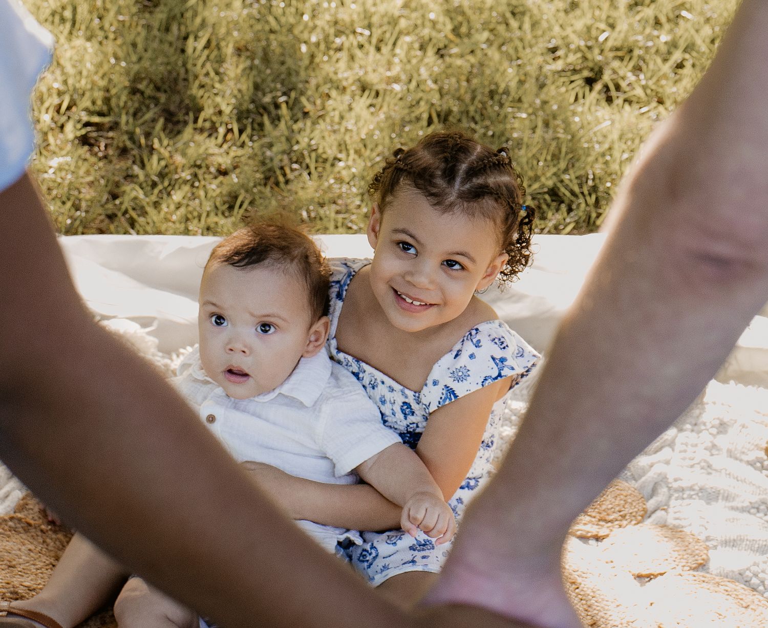 Two young siblings, a smiling toddler girl and baby boy, lying on a blanket outdoors while adults stand nearby.