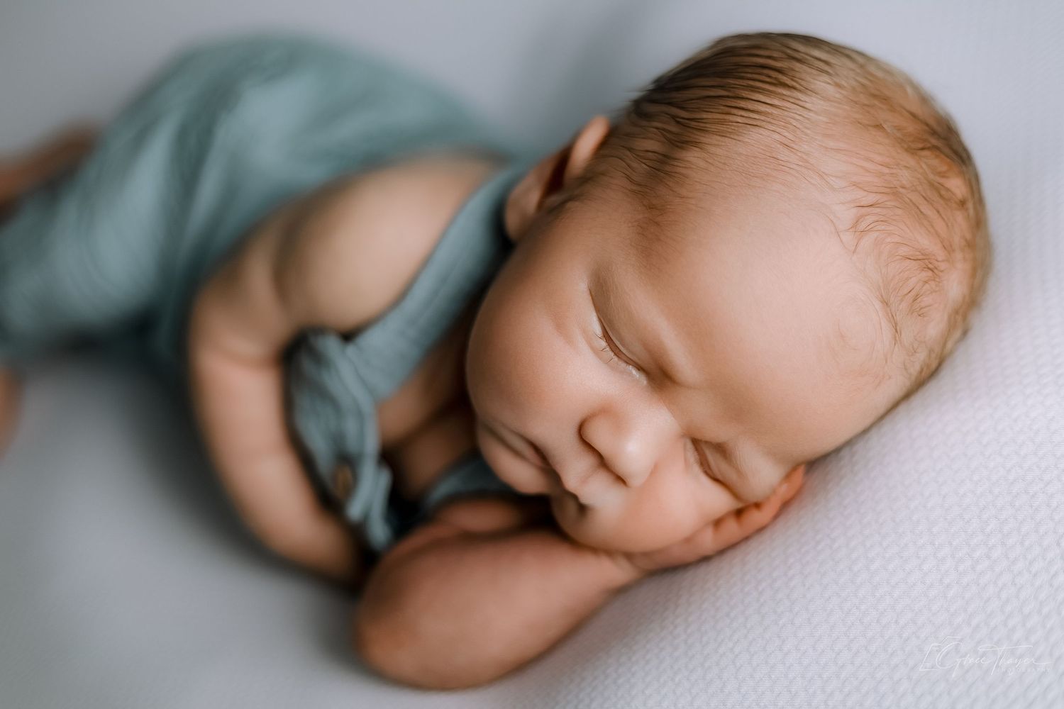 Peaceful newborn baby sleeping in a teal romper on a white blanket, captured in a soft-light portrait photo.