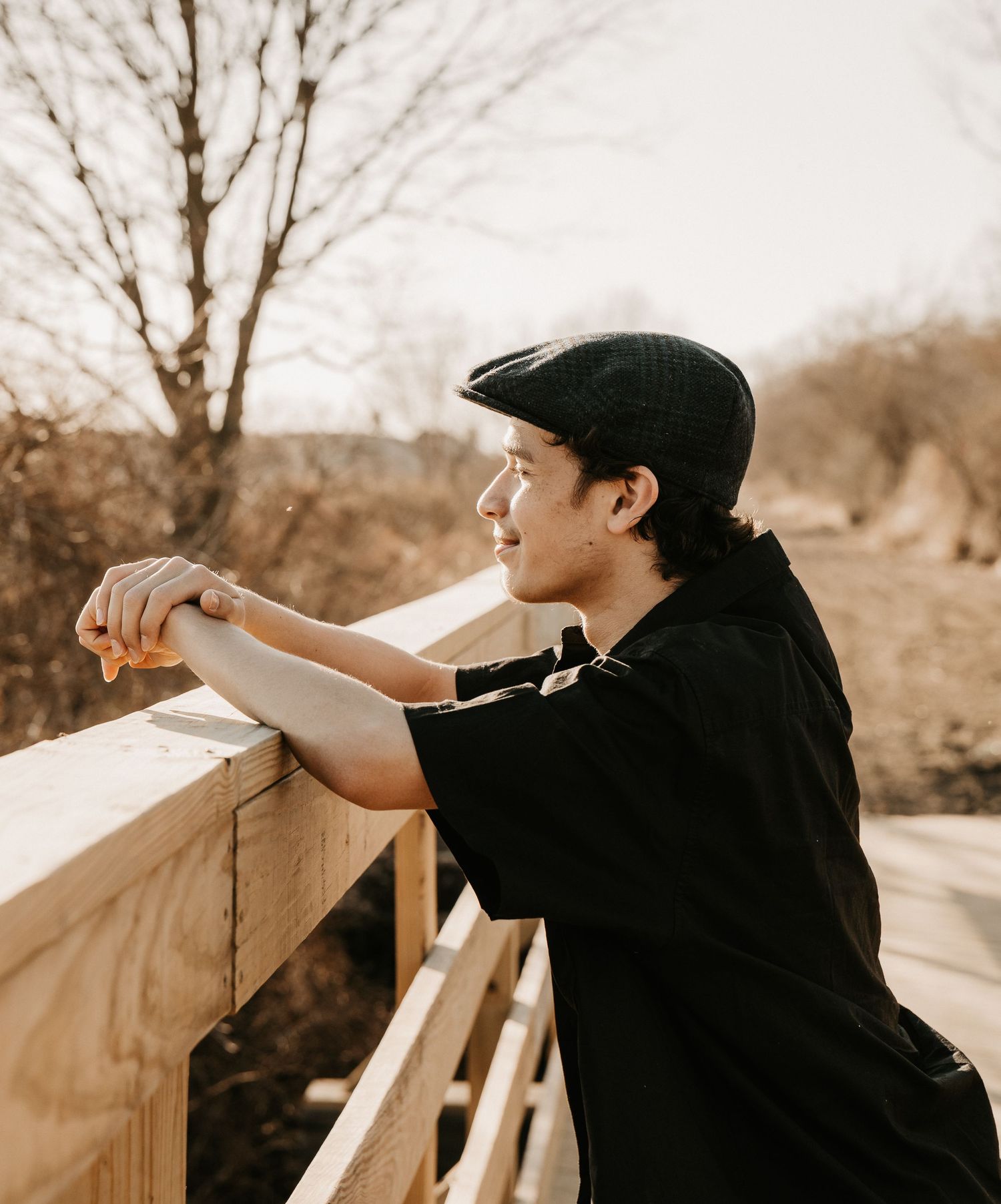 Young man in black outfit and flat cap leaning on wooden railing, smiling outdoors in warm natural light.