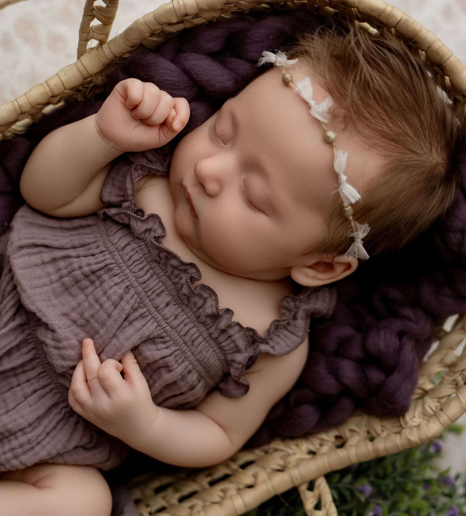 Sleeping newborn baby in a wicker basket with purple yarn, wearing a floral headband and mauve romper.