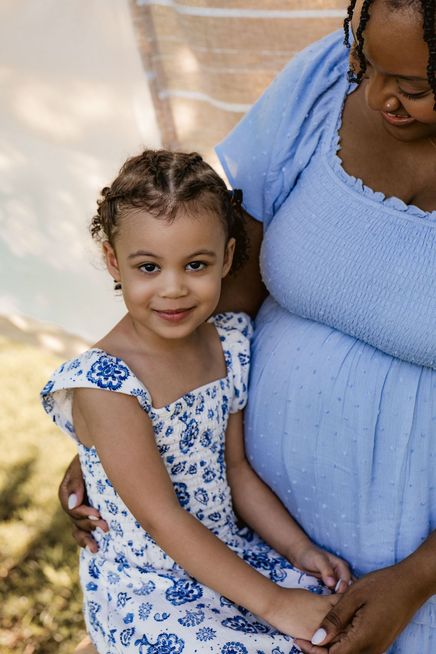 A young girl in a blue floral dress smiles while hugging her pregnant mothers baby bump outdoors.