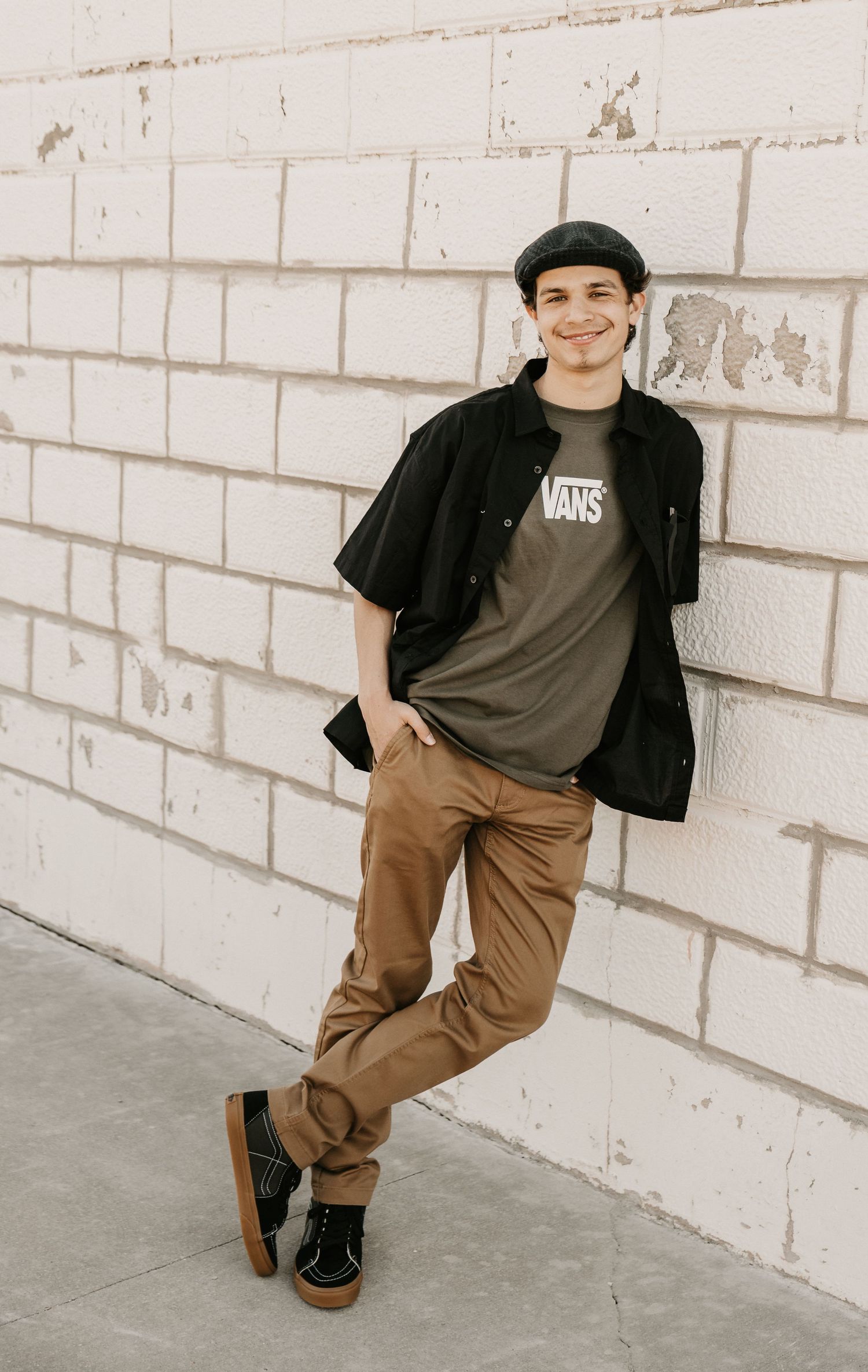 Young man in Vans tee, black overshirt, tan chinos, and beret leaning against white brick wall smiling.