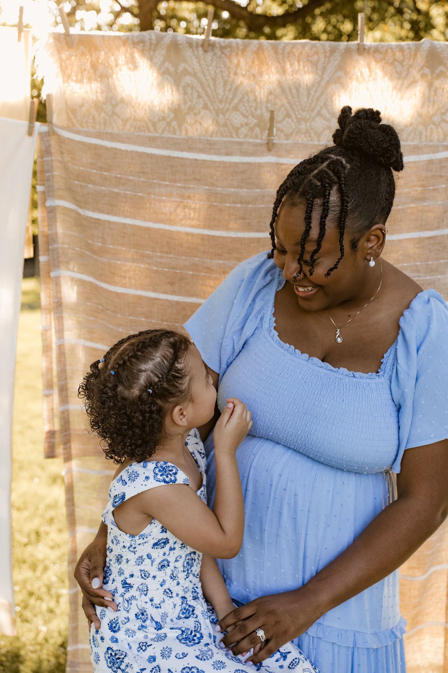 A pregnant woman in a blue dress shares a tender moment with her young daughter outdoors.