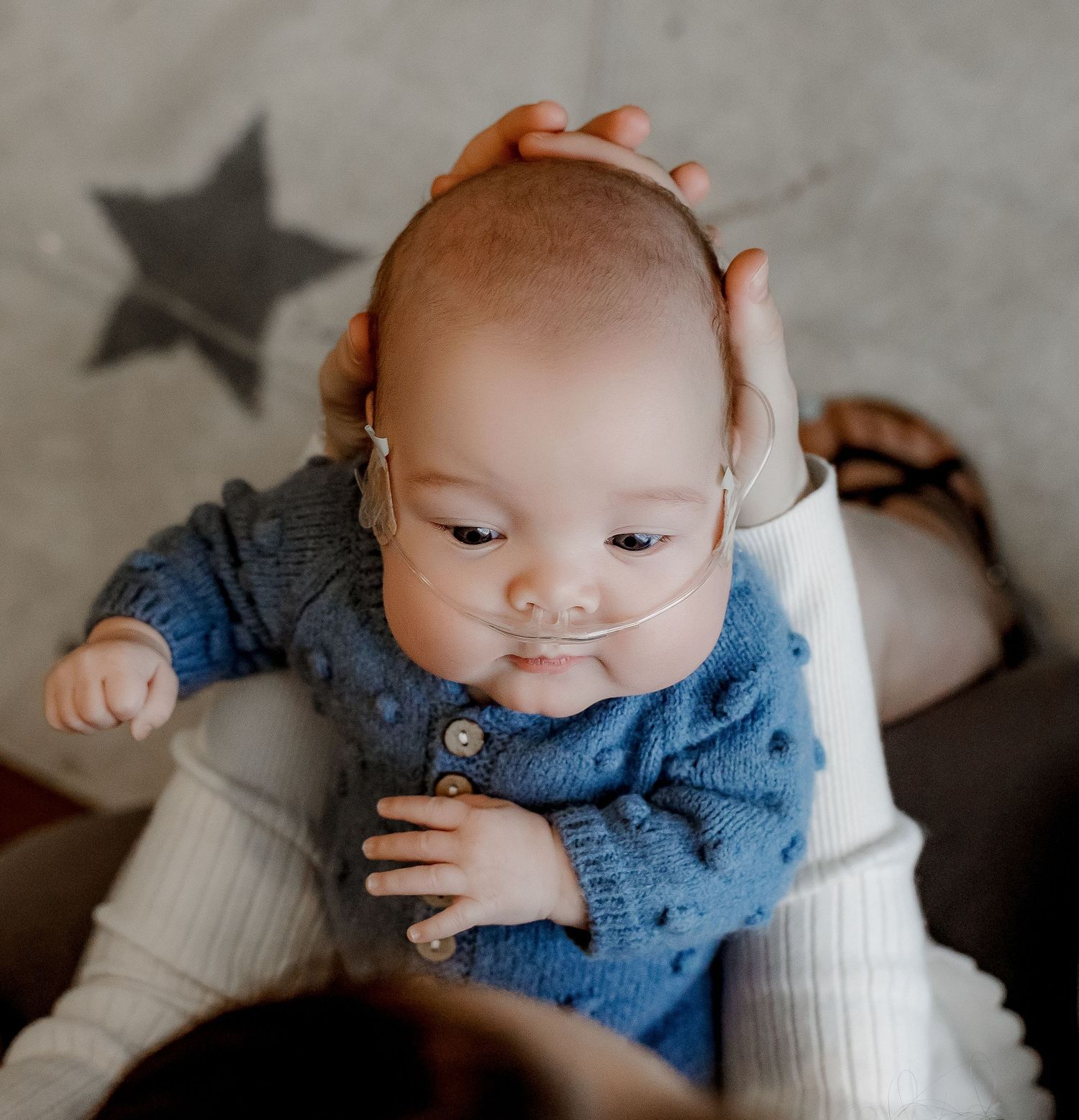 Infant in blue knit cardigan with nasal cannula held gently by caregiver on star-patterned background.
