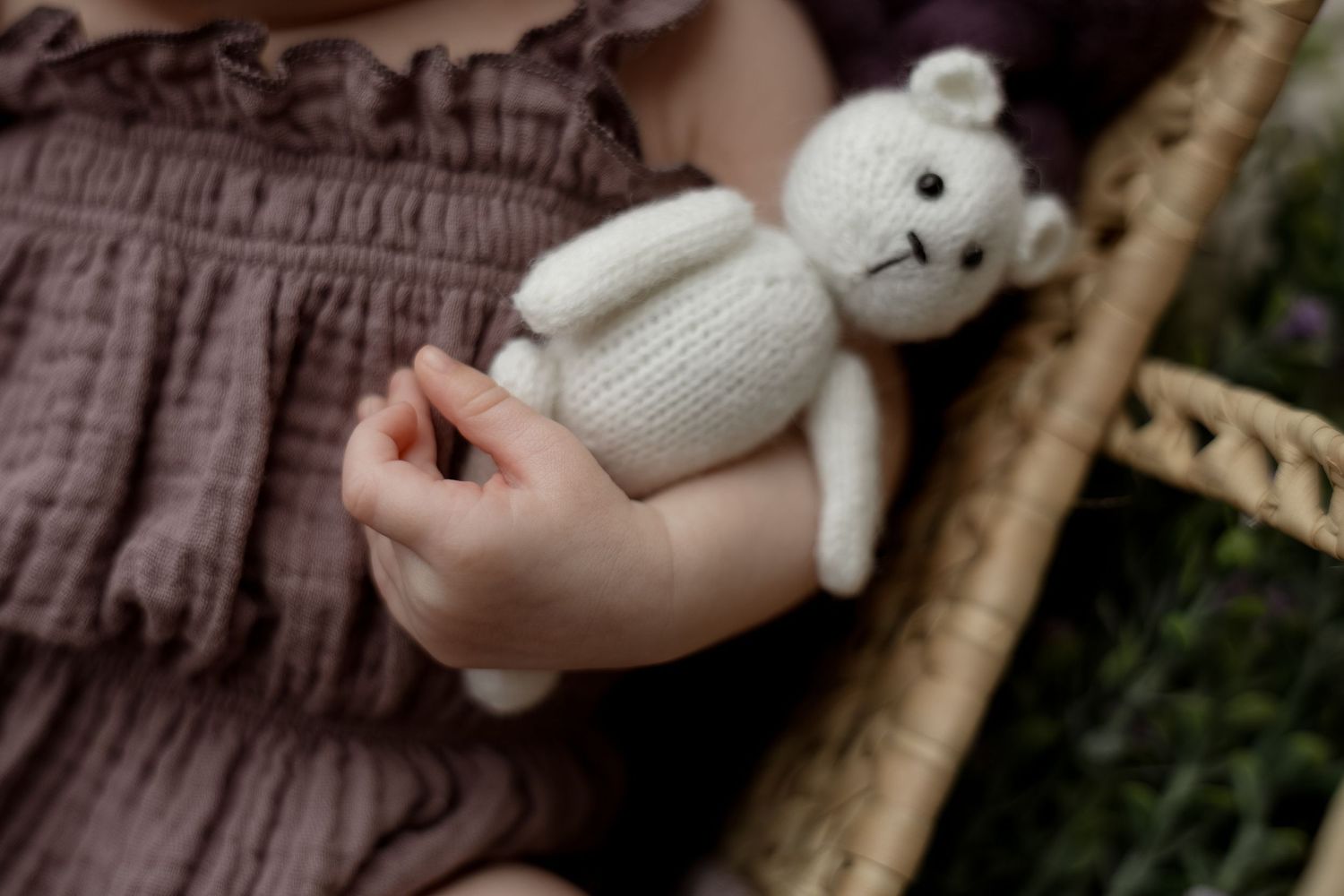 Baby holding a small knitted white teddy bear toy while sitting in a wicker basket wearing a mauve dress.