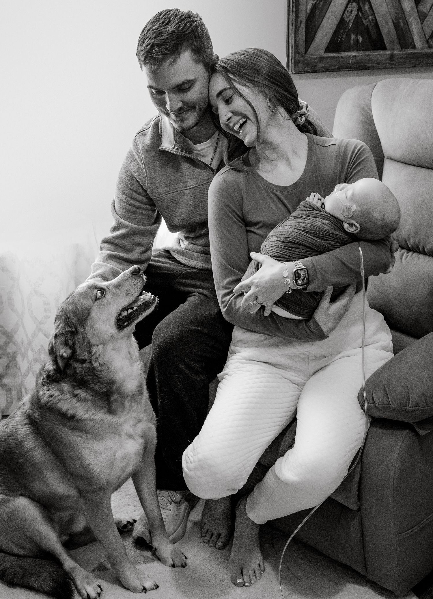Black and white photo of a couple smiling at their newborn baby while a dog looks up at them in a cozy living room.