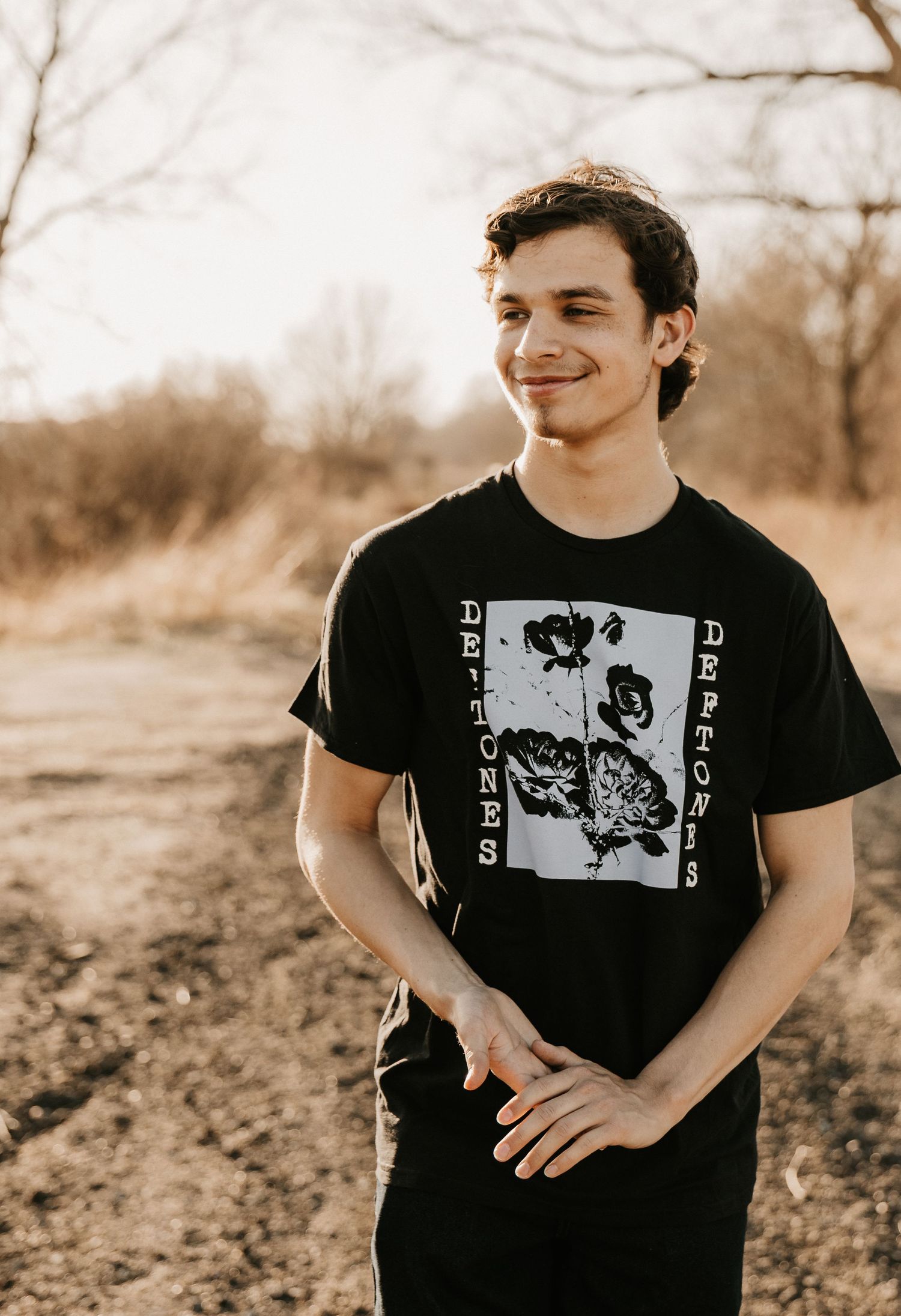 Young man smiling in a black Deftones band t-shirt, standing outdoors in a natural setting with bare trees.