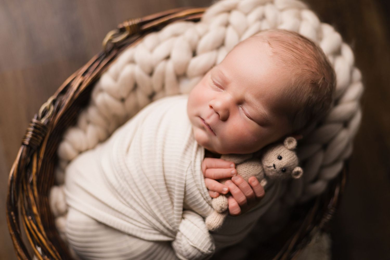 Sleeping newborn wrapped in white swaddle rests in wicker basket with knitted blanket and small stuffed toy.