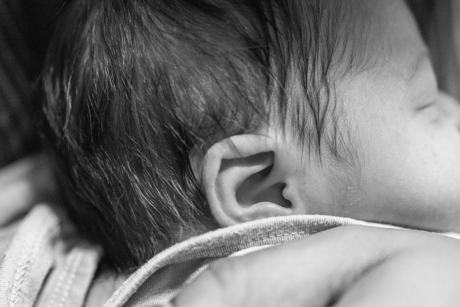 Close-up black and white photo of a newborn baby's ear and dark hair.