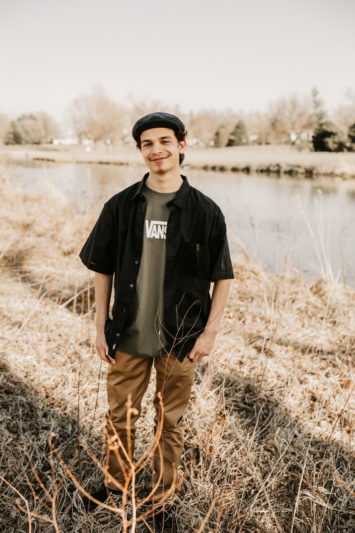 Teen boy in Vans tee and black shirt smiling by a pond in dry grass field.