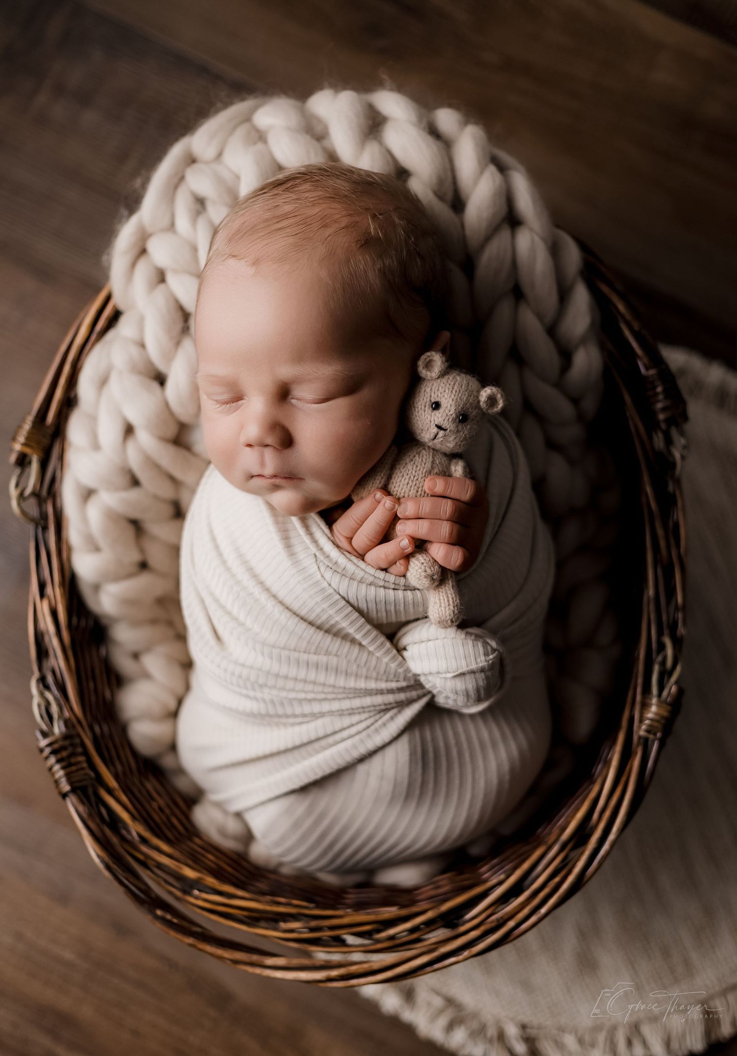 Newborn baby swaddled in cream wrap holding a tiny teddy bear, sleeping peacefully in a wicker basket with chunky knit blanket.