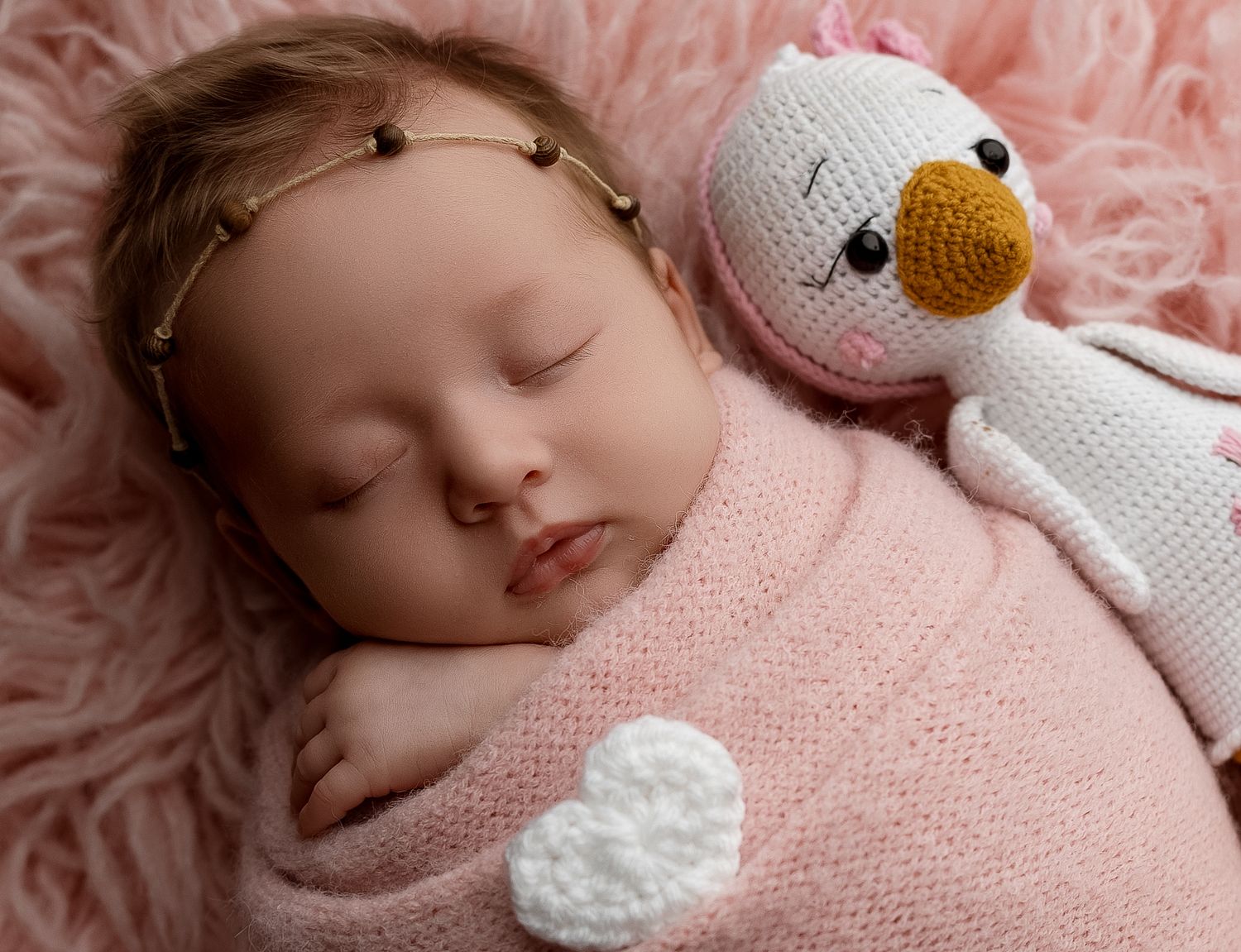 Sleeping newborn baby wrapped in pink knit blanket with crocheted duck toy on fluffy pink background.