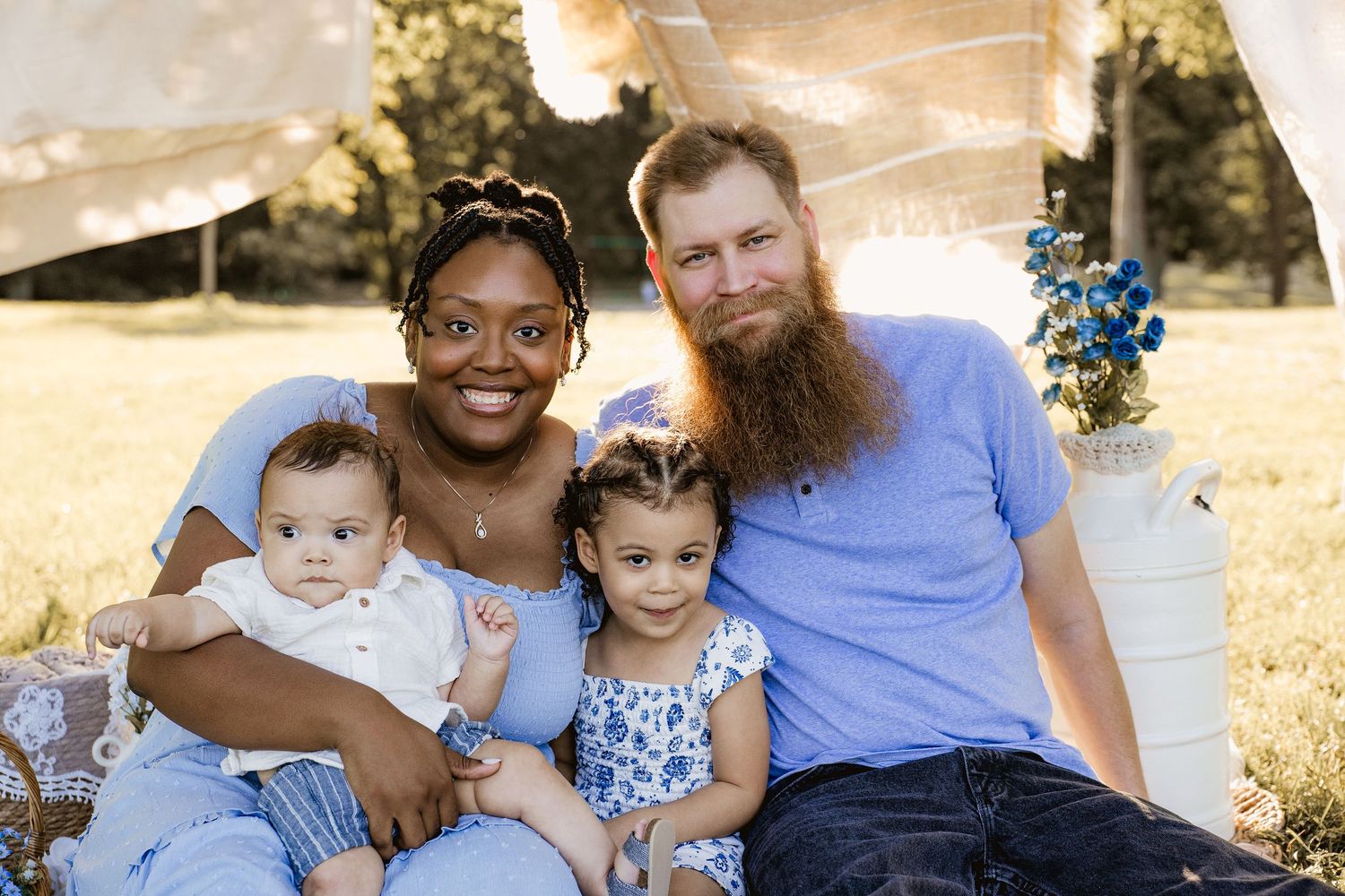 Interracial family portrait outdoors, parents with two young children sitting on blanket in sunny park with hanging fabric backdrop.
