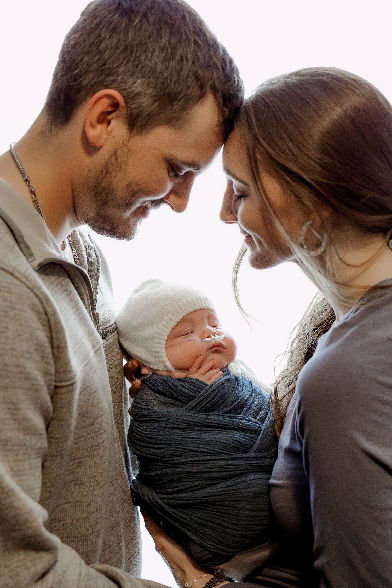 Parents tenderly touch foreheads while holding their swaddled newborn baby in a white knit hat.