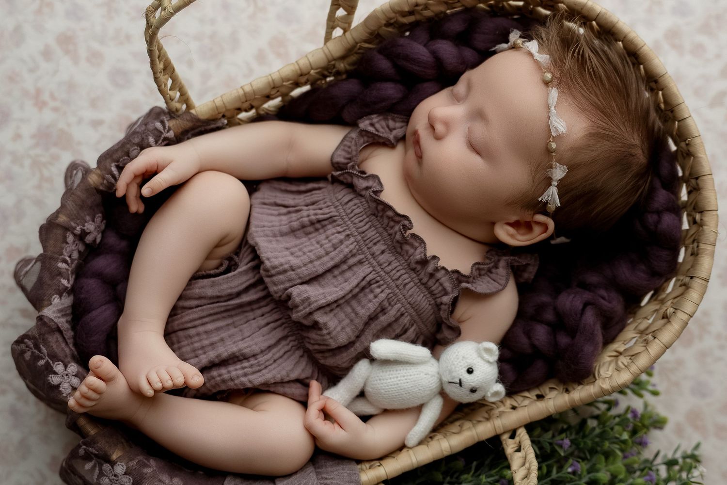 Sleeping newborn baby in a wicker basket with purple wool, wearing a mauve romper, holding a white teddy bear.