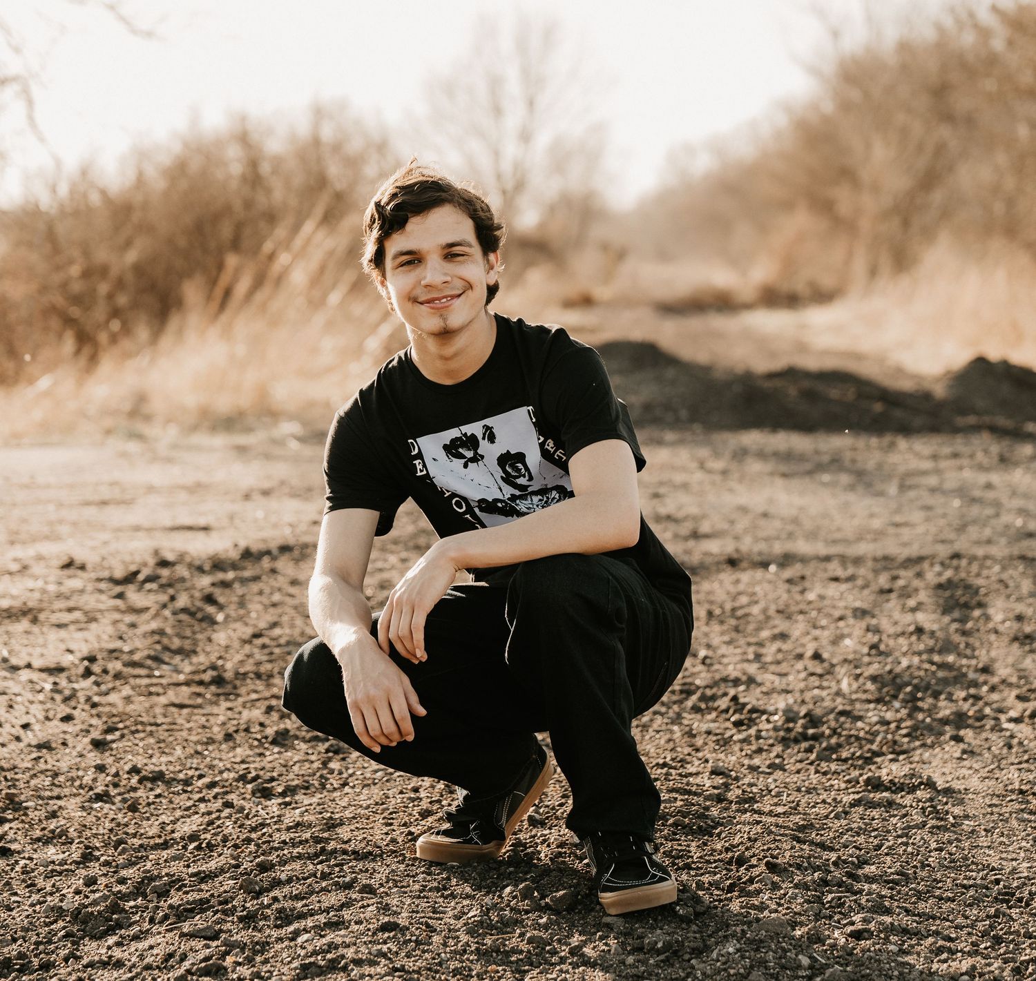 Young man in black graphic tee crouching outdoors on dirt ground with dry brush and trees in background.