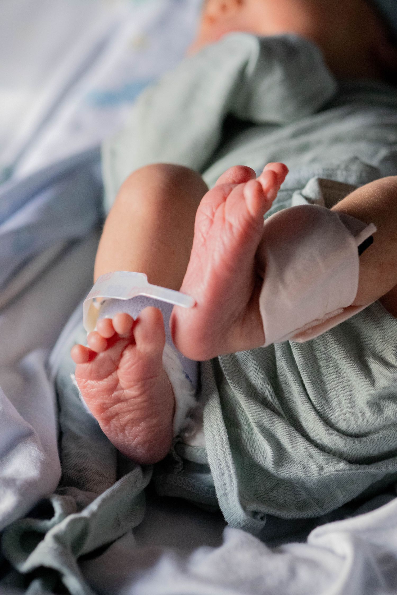 Newborn baby feet with hospital ID bracelet, tiny wrinkled toes visible in soft green blanket.