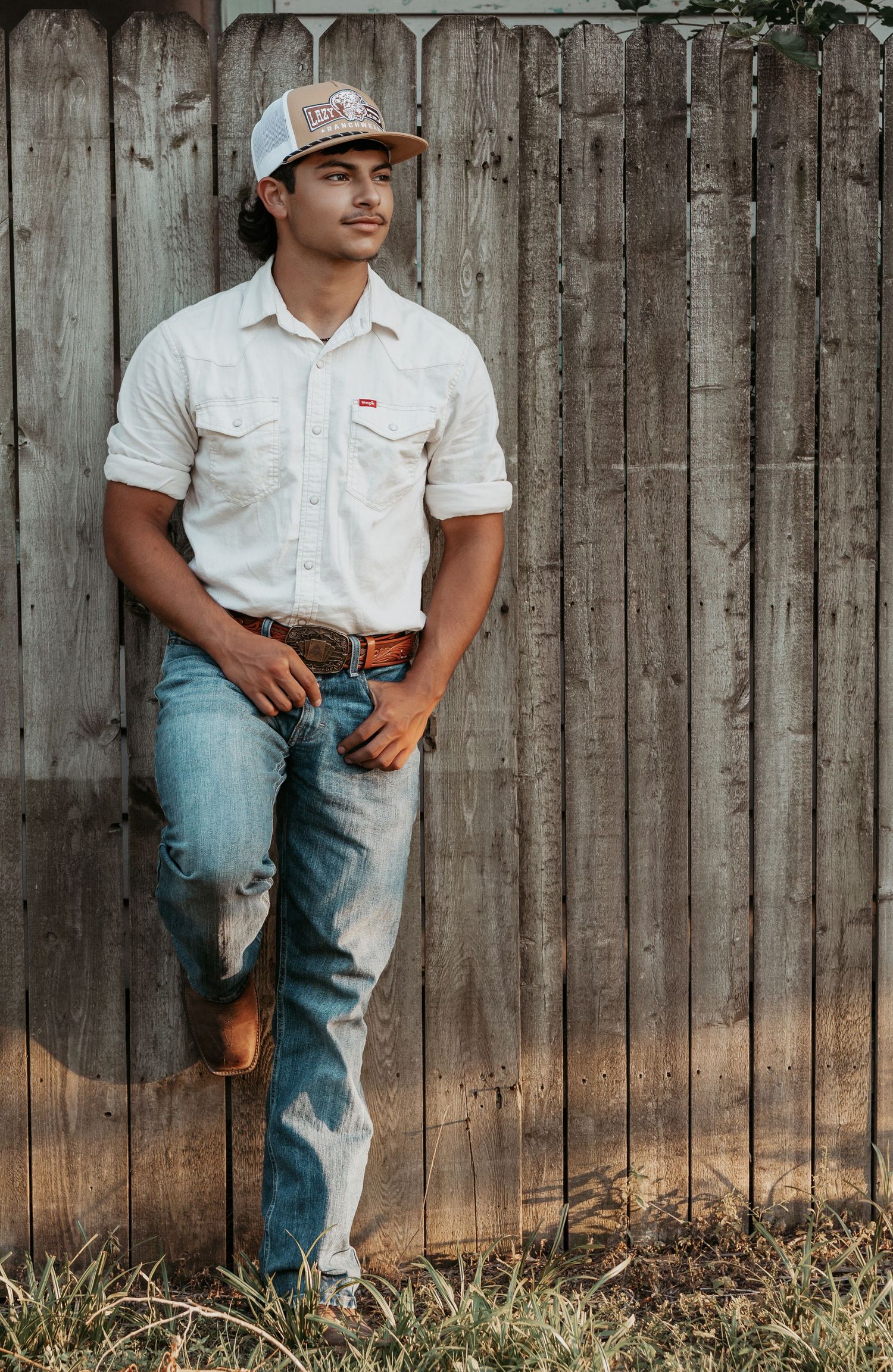 A young man in a white Levi's shirt, jeans, and trucker hat leans against a rustic wooden fence.