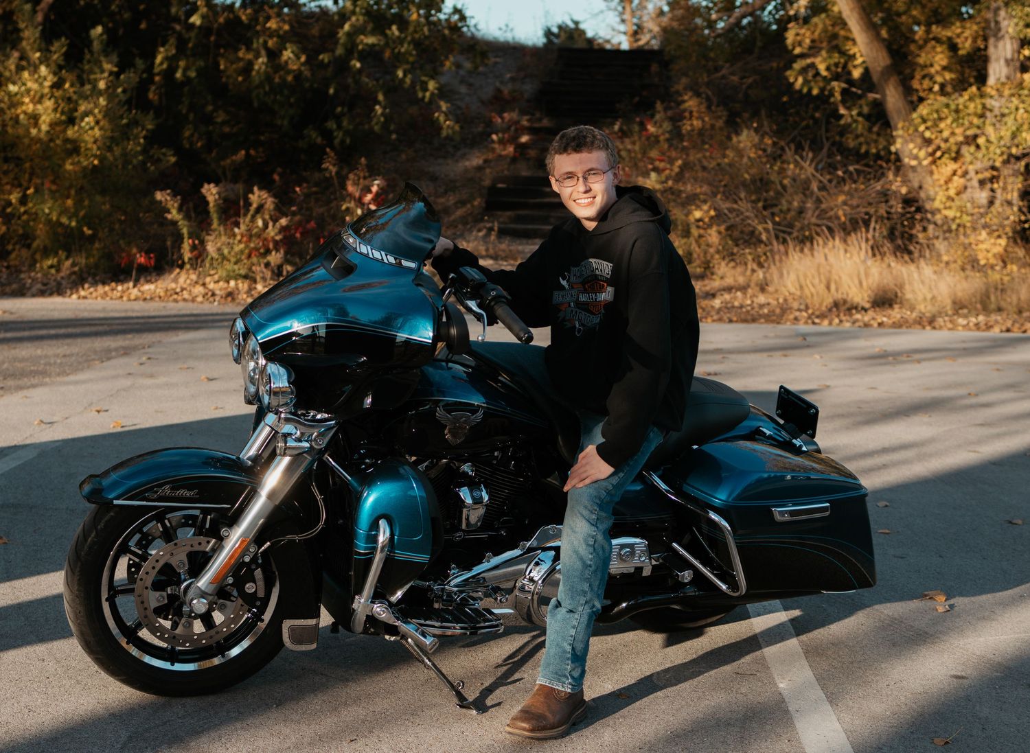 Man in black jacket posing with a teal Harley-Davidson motorcycle on a road surrounded by autumn trees.