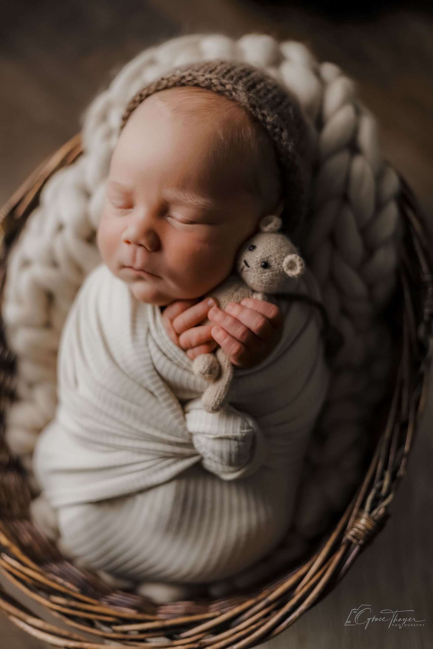Newborn baby swaddled in white wrap, sleeping in a wicker basket with chunky knit blanket, holding a tiny teddy bear.
