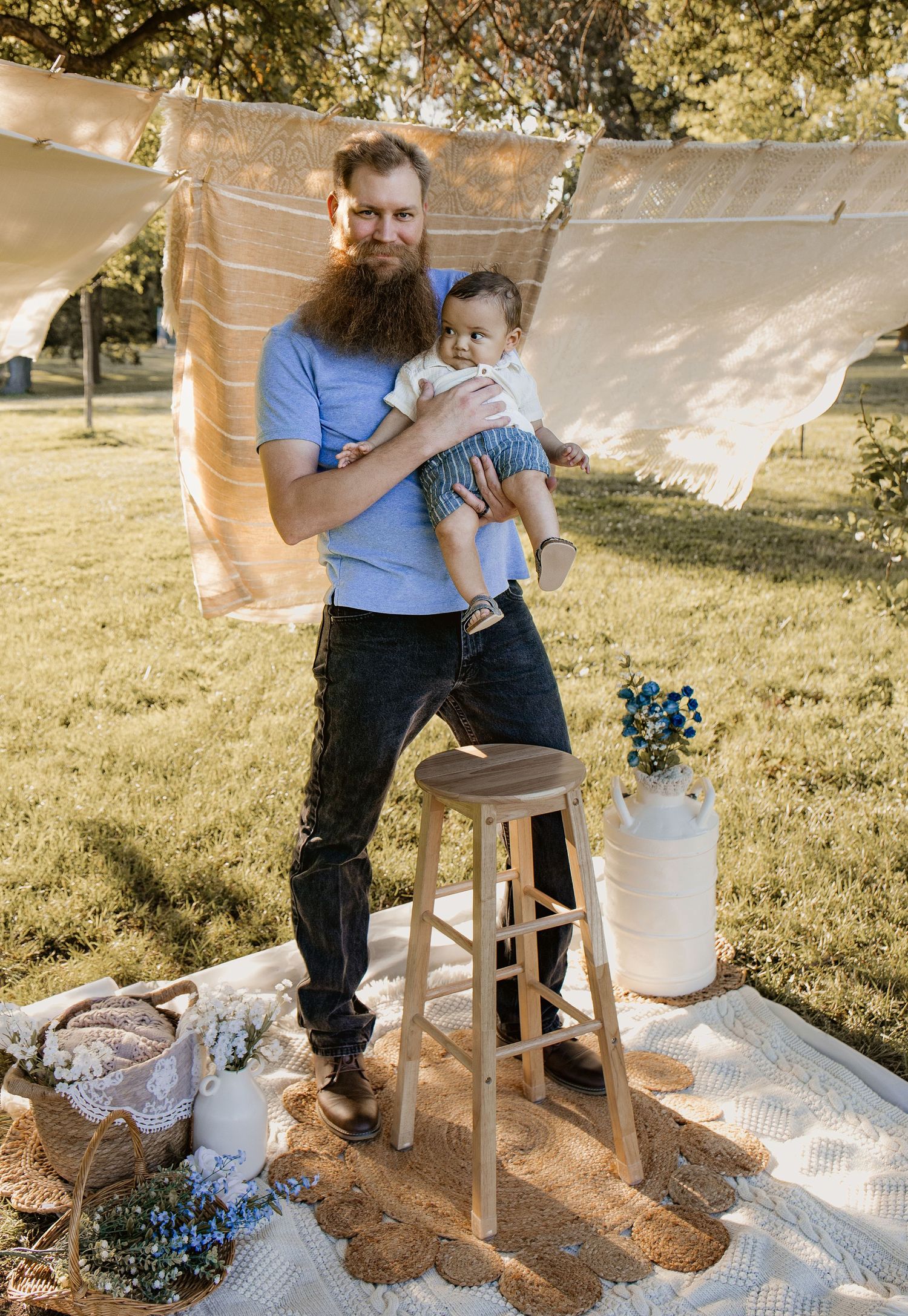 Bearded red-haired man holding infant baby outdoors on blanket with wooden stool and floral decorations in sunny park.