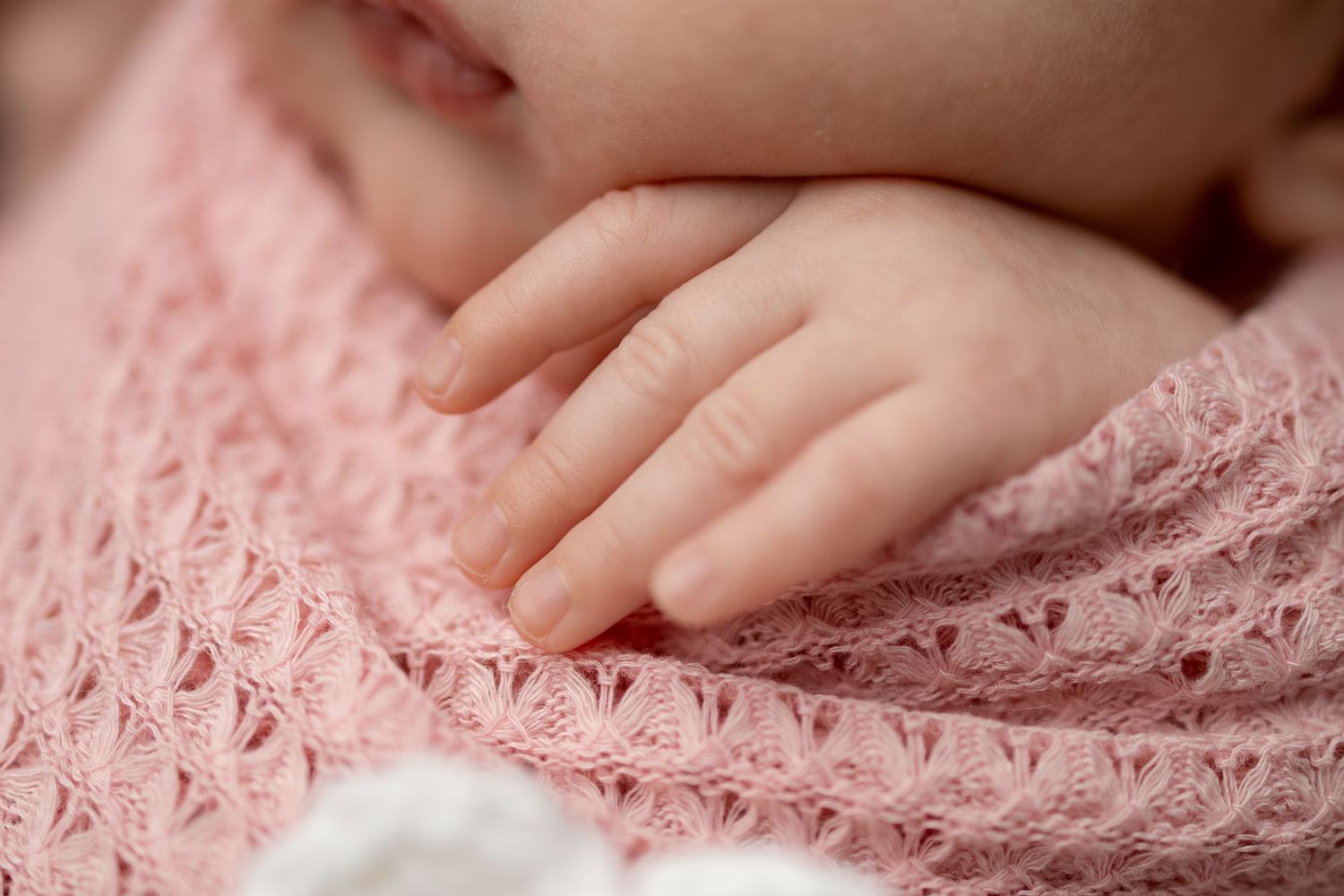 Close-up of a newborn baby's tiny hand resting on a soft pink lace knit wrap blanket.