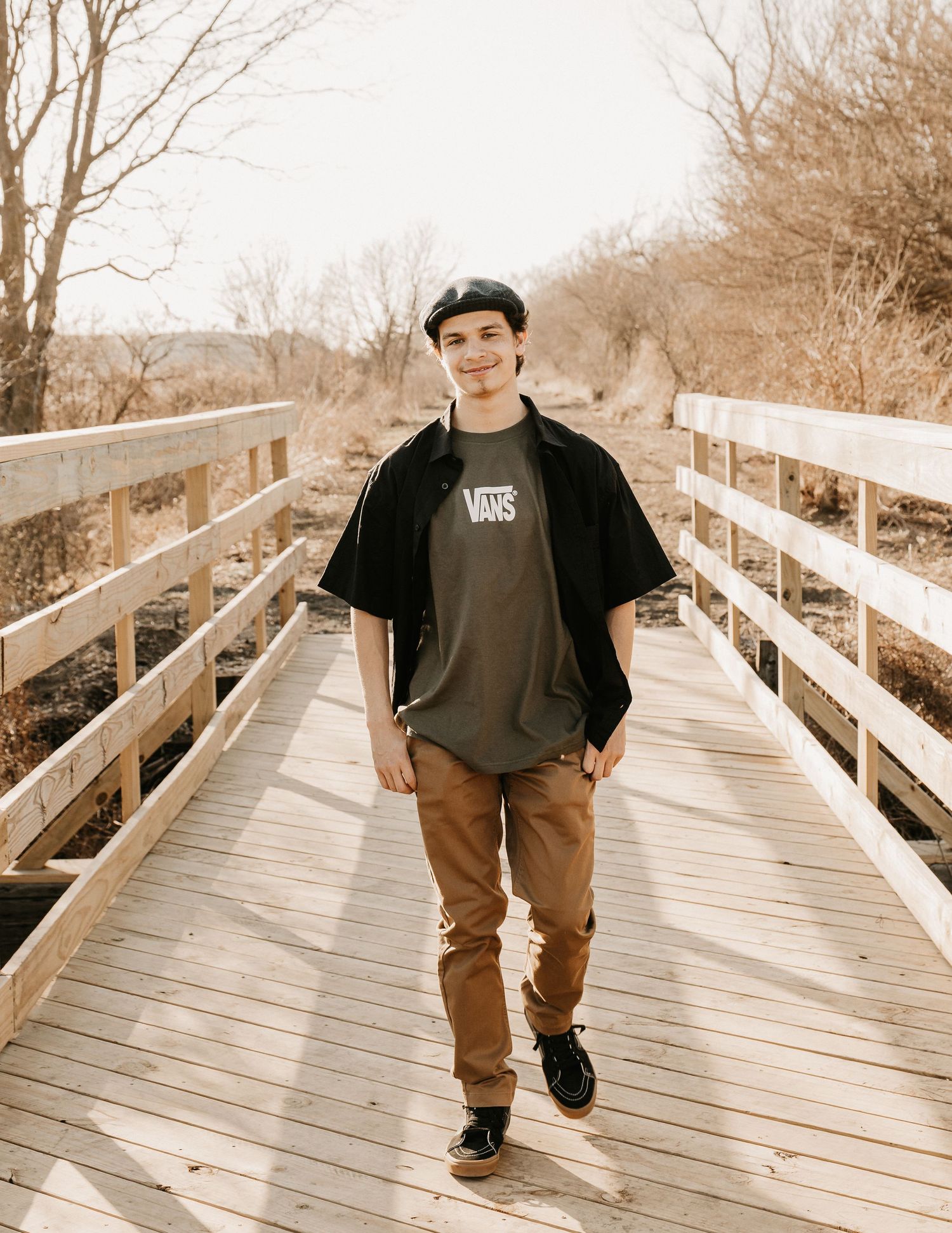 Teen boy in Vans tee and cap walks on wooden bridge surrounded by bare trees in natural outdoor setting.