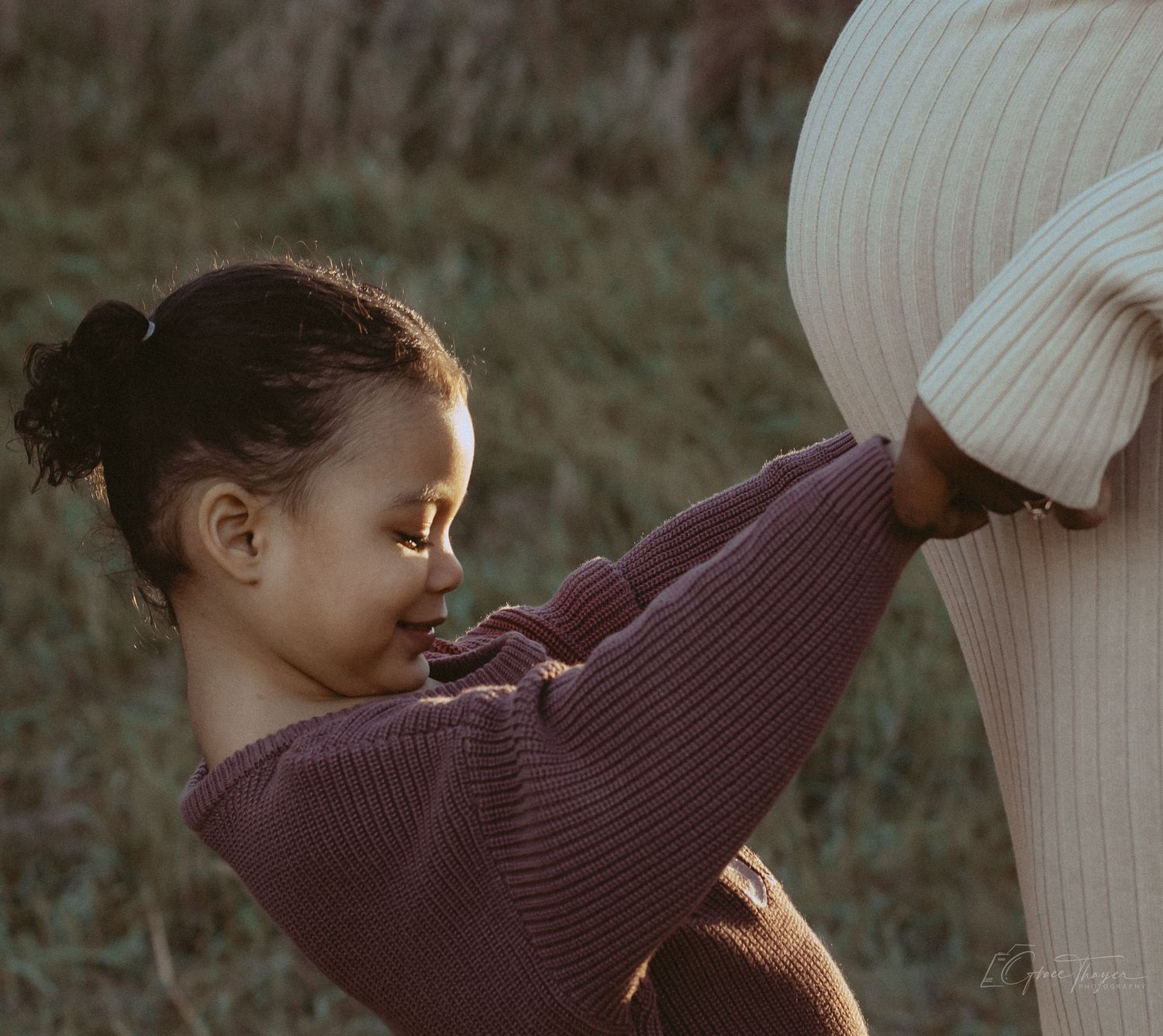 A young girl in a purple sweater smiles while hugging a pregnant woman's belly outdoors.
